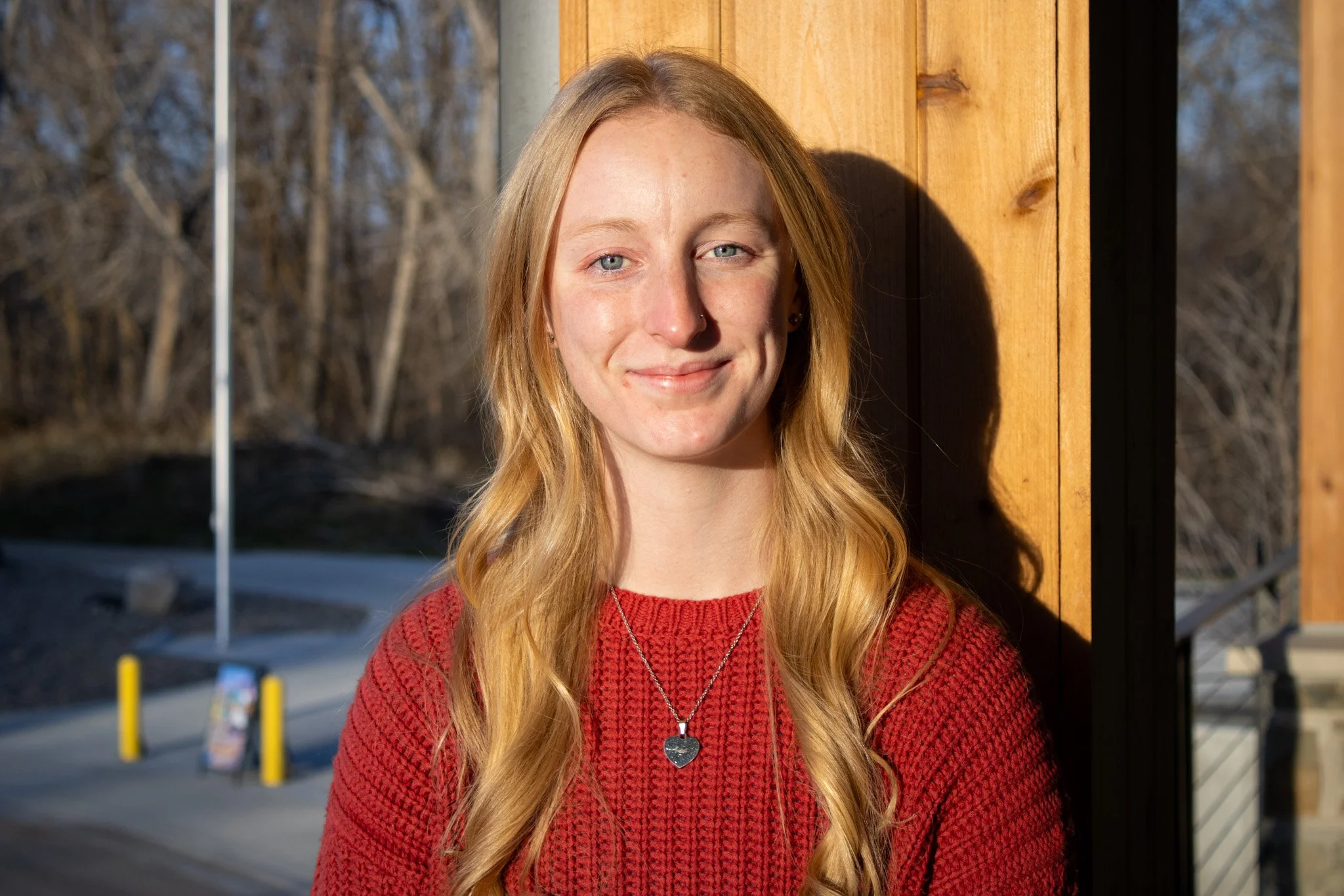 A young woman with long, wavy blonde hair is smiling and standing outdoors, leaning against a wooden wall. She is wearing a red sweater and a silver heart-shaped necklace. The background features trees and a bright, sunny day.