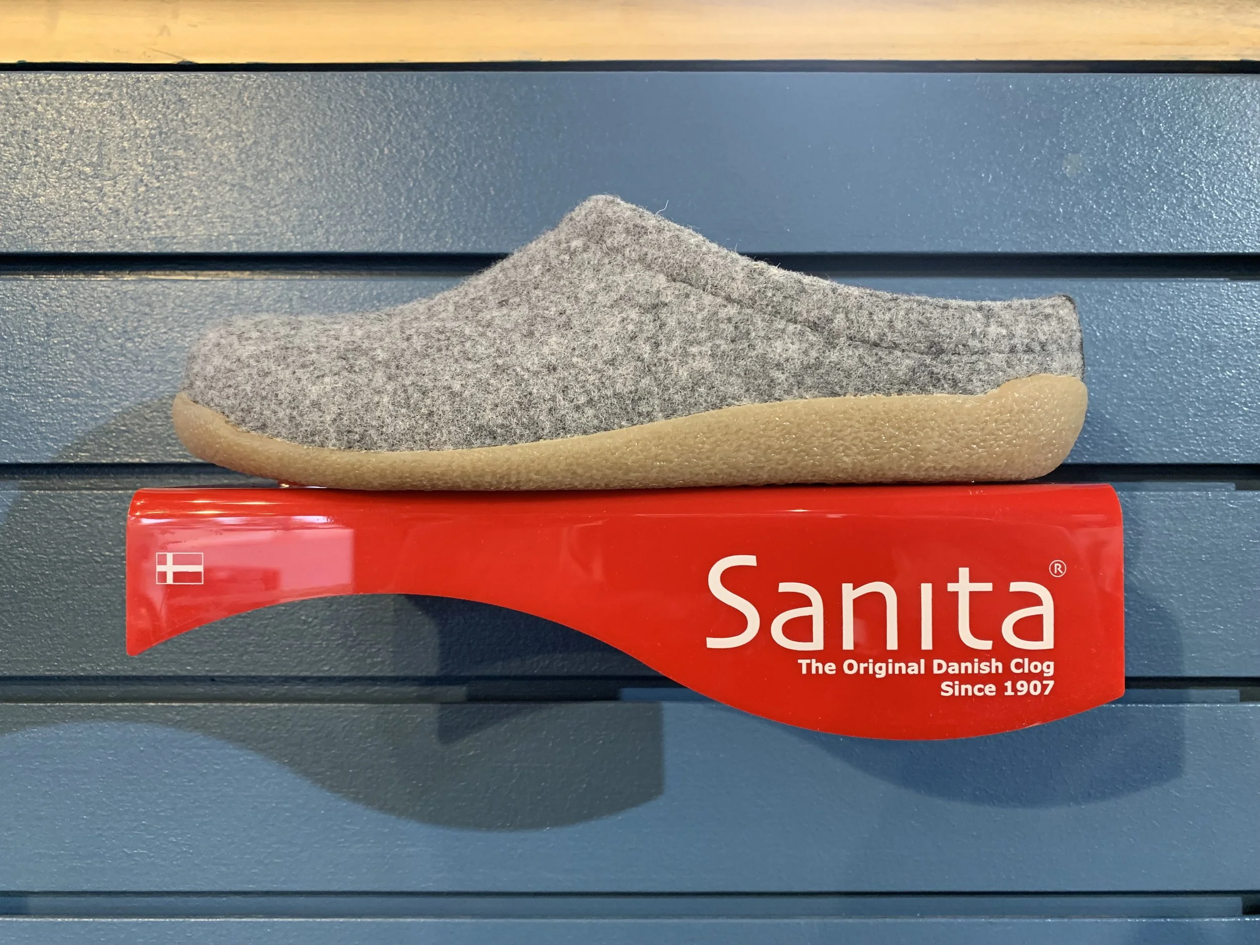 Gray felt slipper placed on top of a red box labeled 'Sanita - The Original Danish Clog Since 1907' with a Danish flag icon, against a textured blue background.