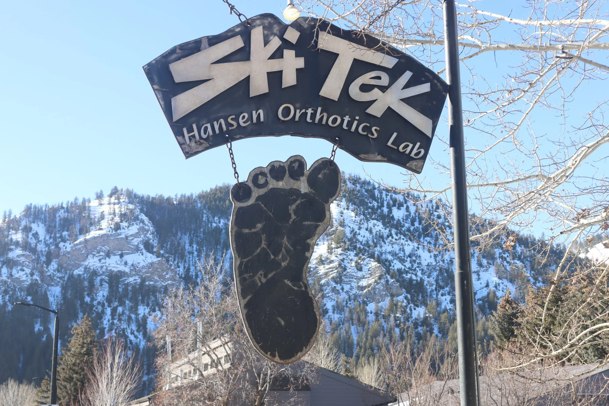 Sign for Hansen Orthotics Lab with a large black footprint hanging below, against a backdrop of snowy mountains and leafless trees on a clear day.