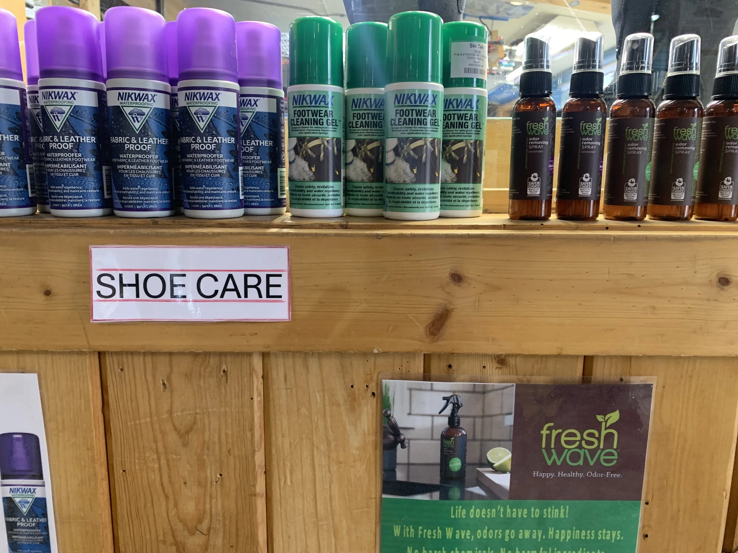 Shoe care products including spray bottles and cans on a wooden shelf with a sign that says 'SHOE CARE' and an advertisement for Fresh Wave odor eliminator spray.