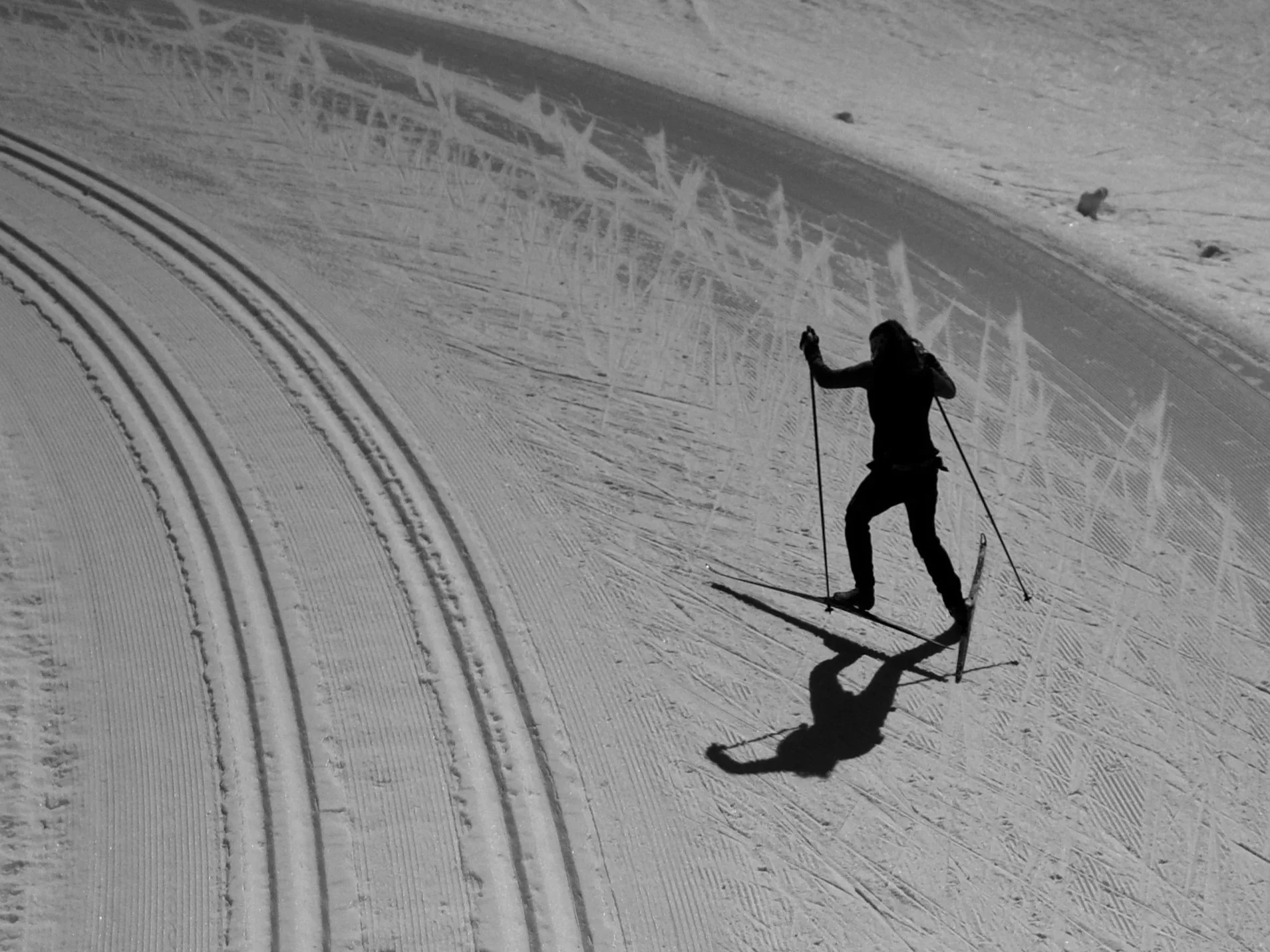 A person skate skiing on a groomed track, casting a shadow on the snow with ski tracks around.