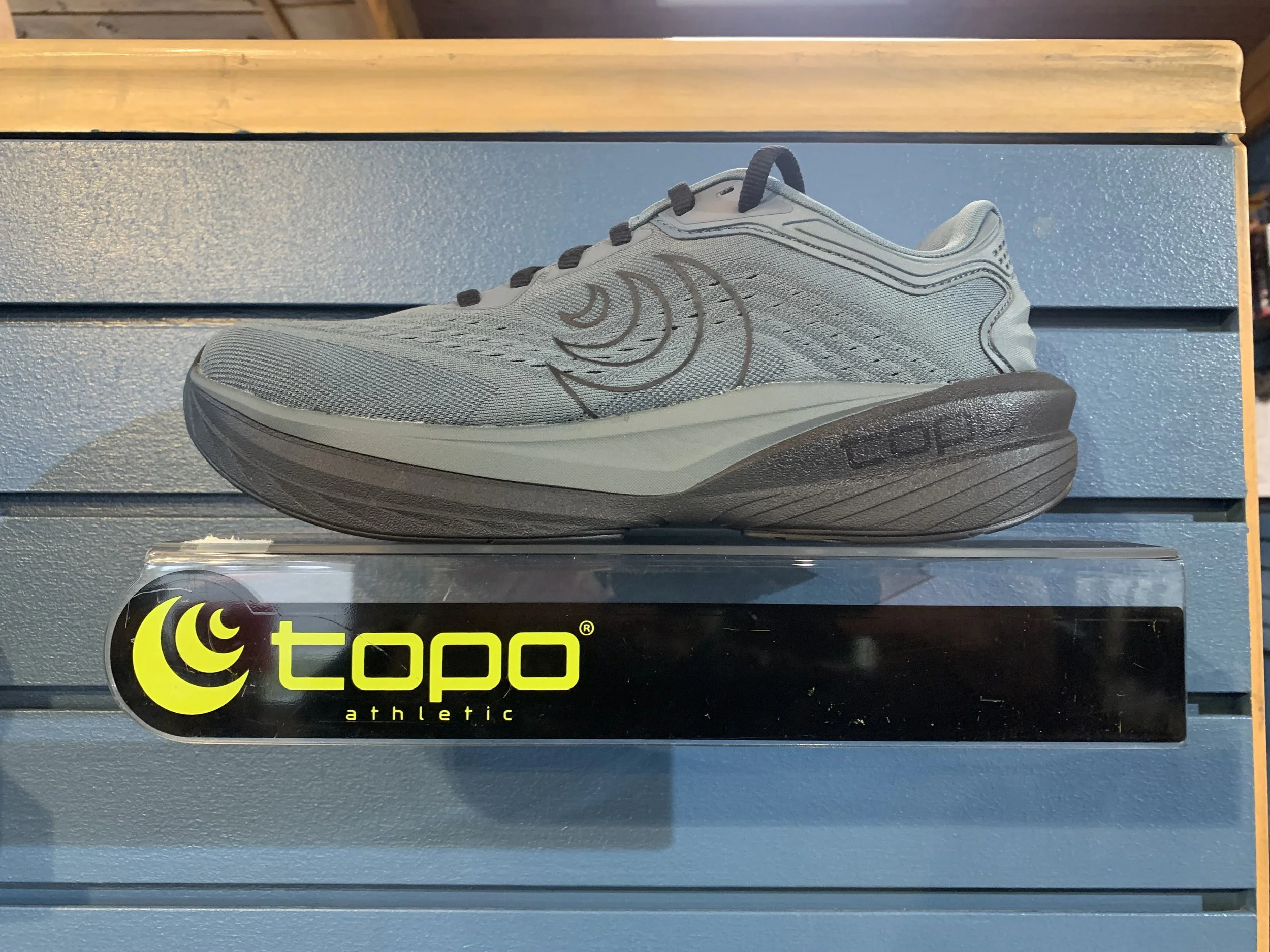 Gray athletic running shoe displayed on a blue slat wall shelf, with a 'topo athletic' logo on a black tag below.