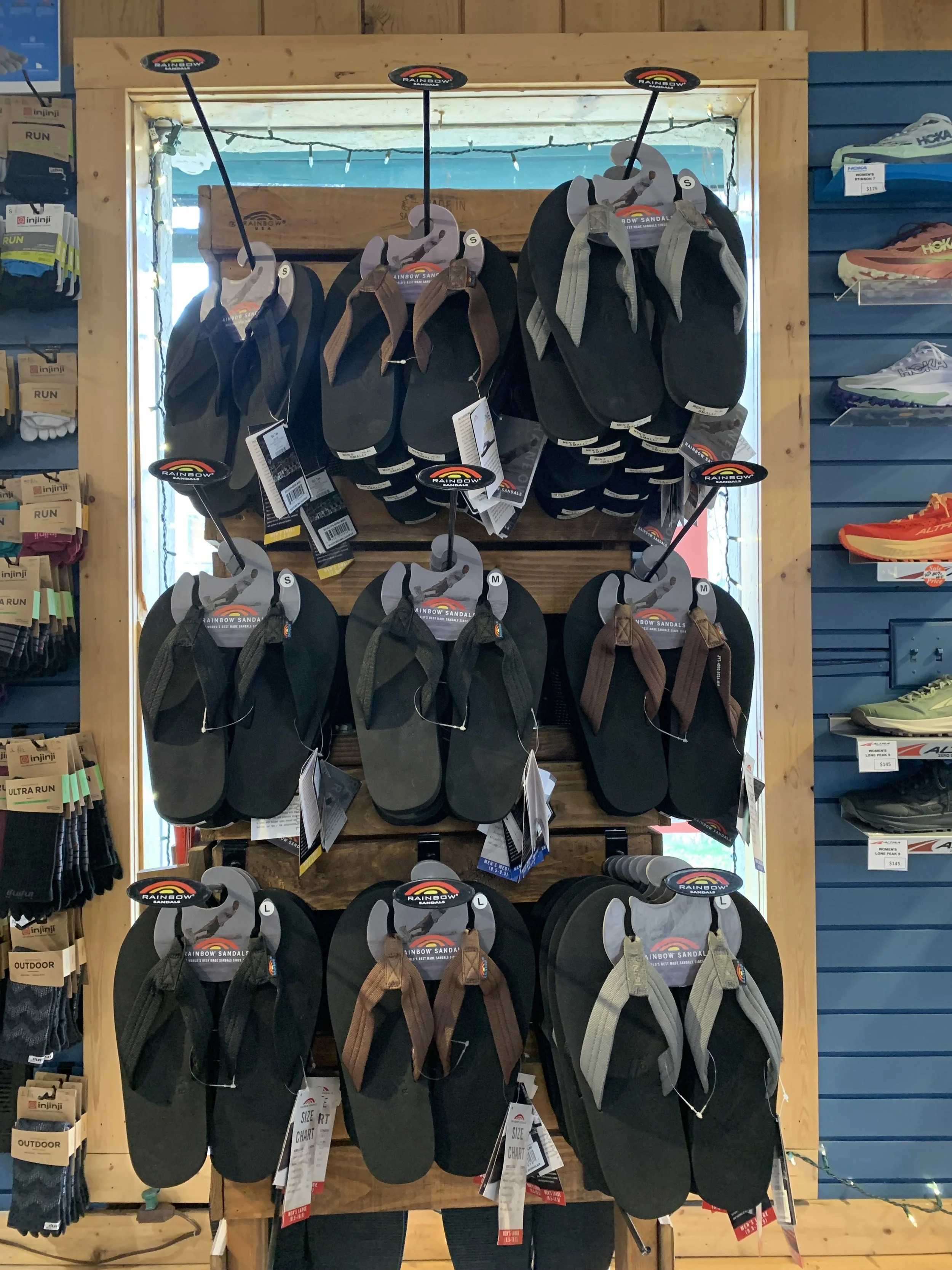 Display of rainbow-colored sandals in various sizes on a wooden store shelf, with tags and hooks for shopping.