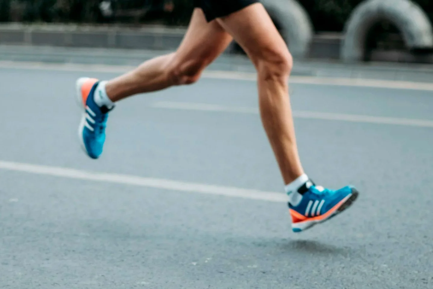 Runner in mid-stride on a paved road, wearing blue running shoes with white stripes, black shorts, and no shirt.