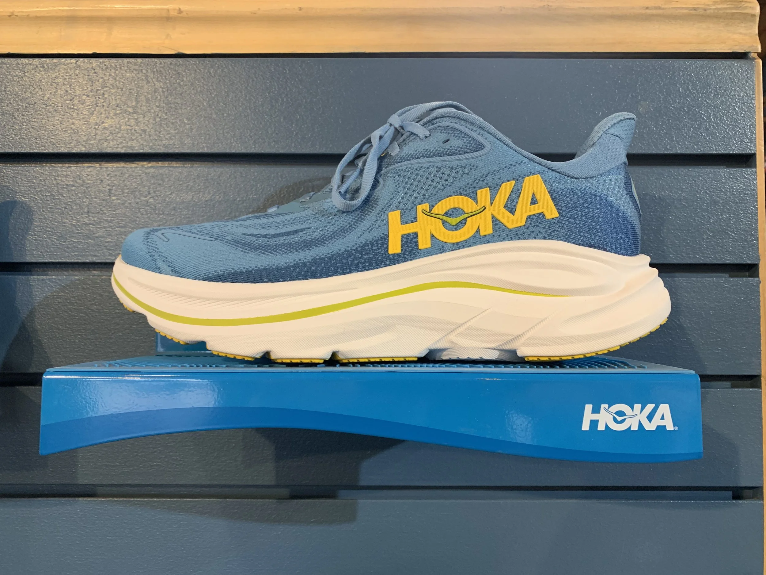 Blue Hoka running shoe with yellow logo, displayed on a blue platform against a gray slatted background.