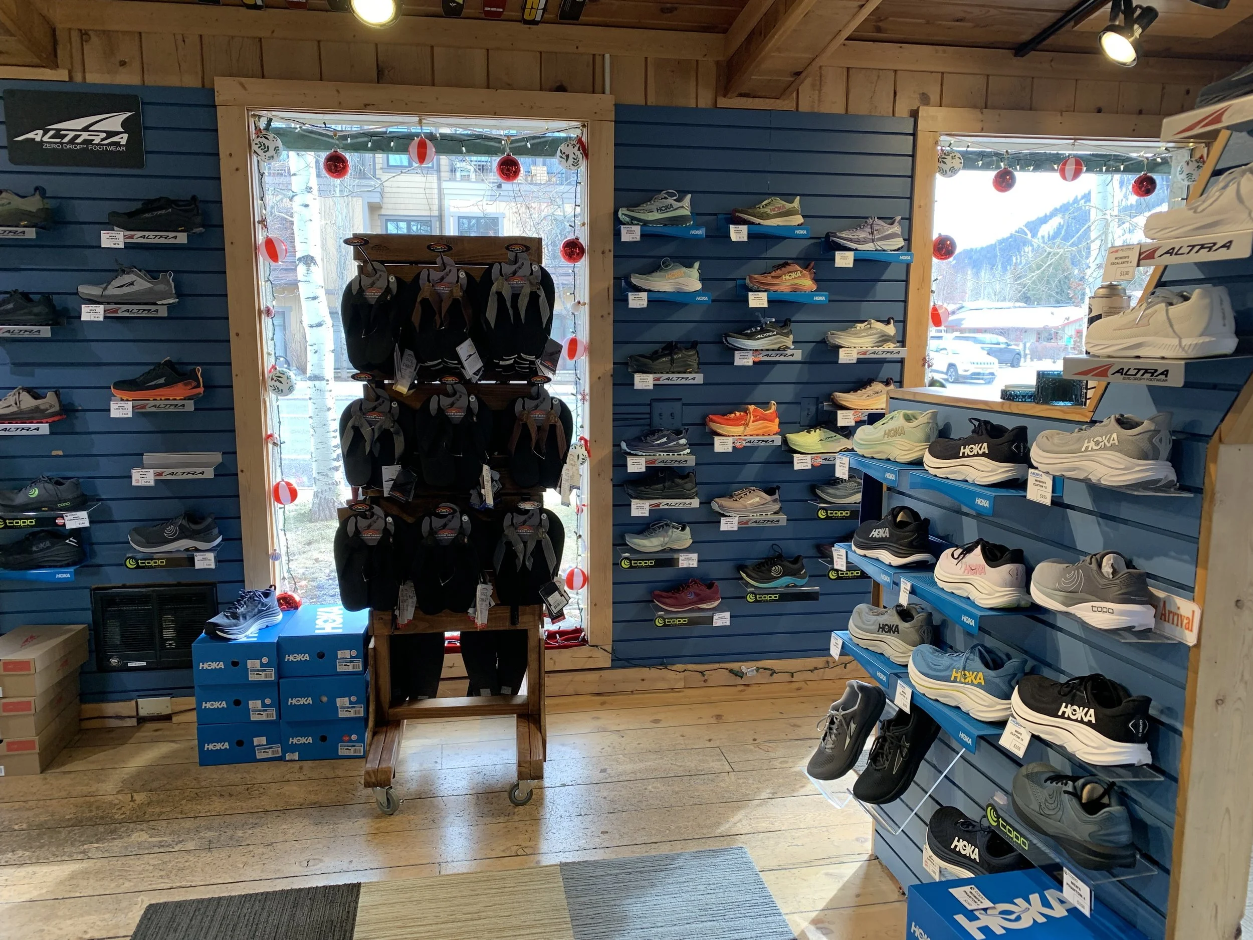 Shoe display in outdoor sports store with Hoka and Altra sneakers, and Rainbow sandals, next to windows decorated with holiday ornaments.