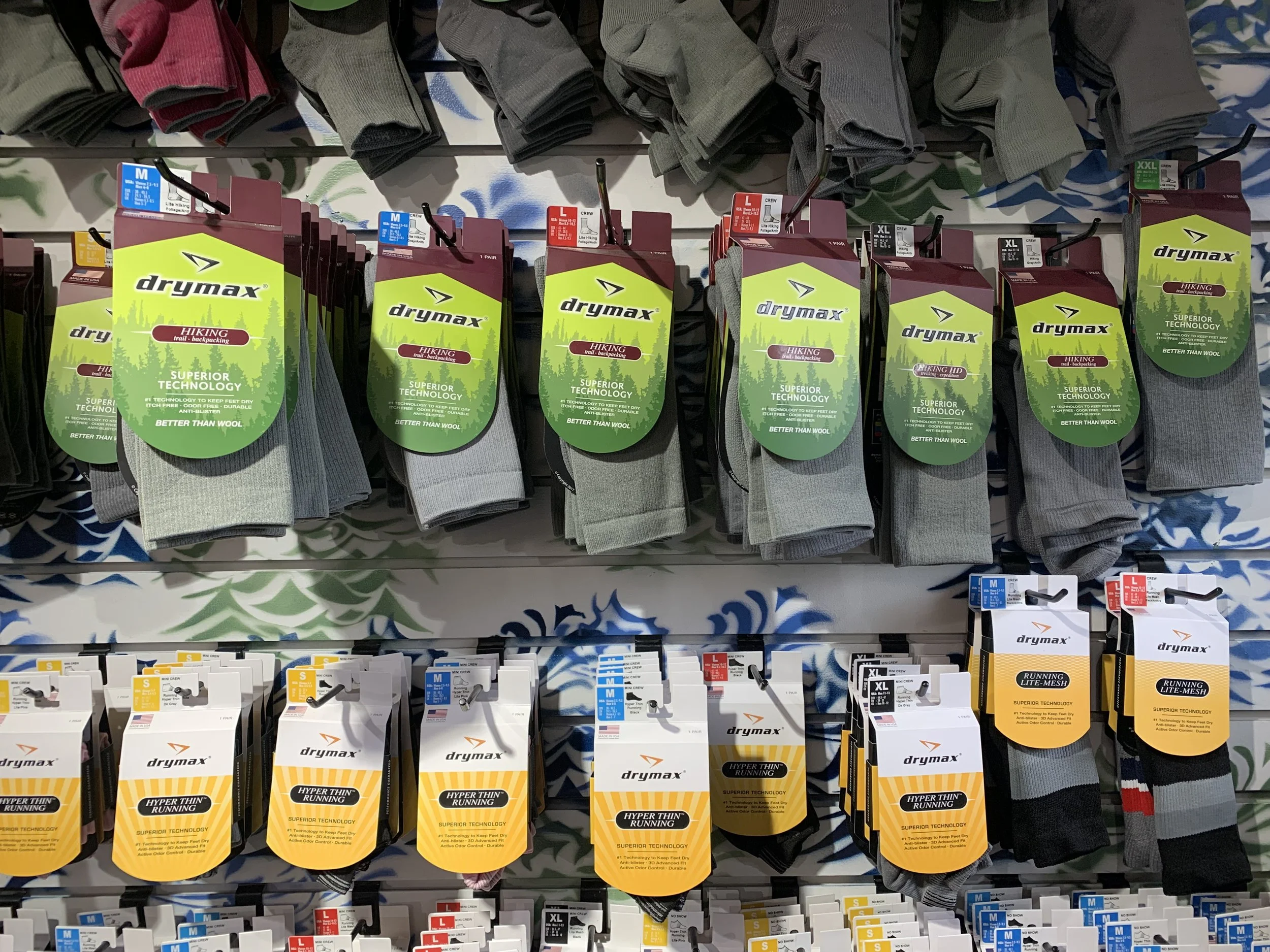 Display of men's socks in a retail store, including gray and black socks in various sizes with labels indicating Drymax brand and features like superior technology and hyper thin running.