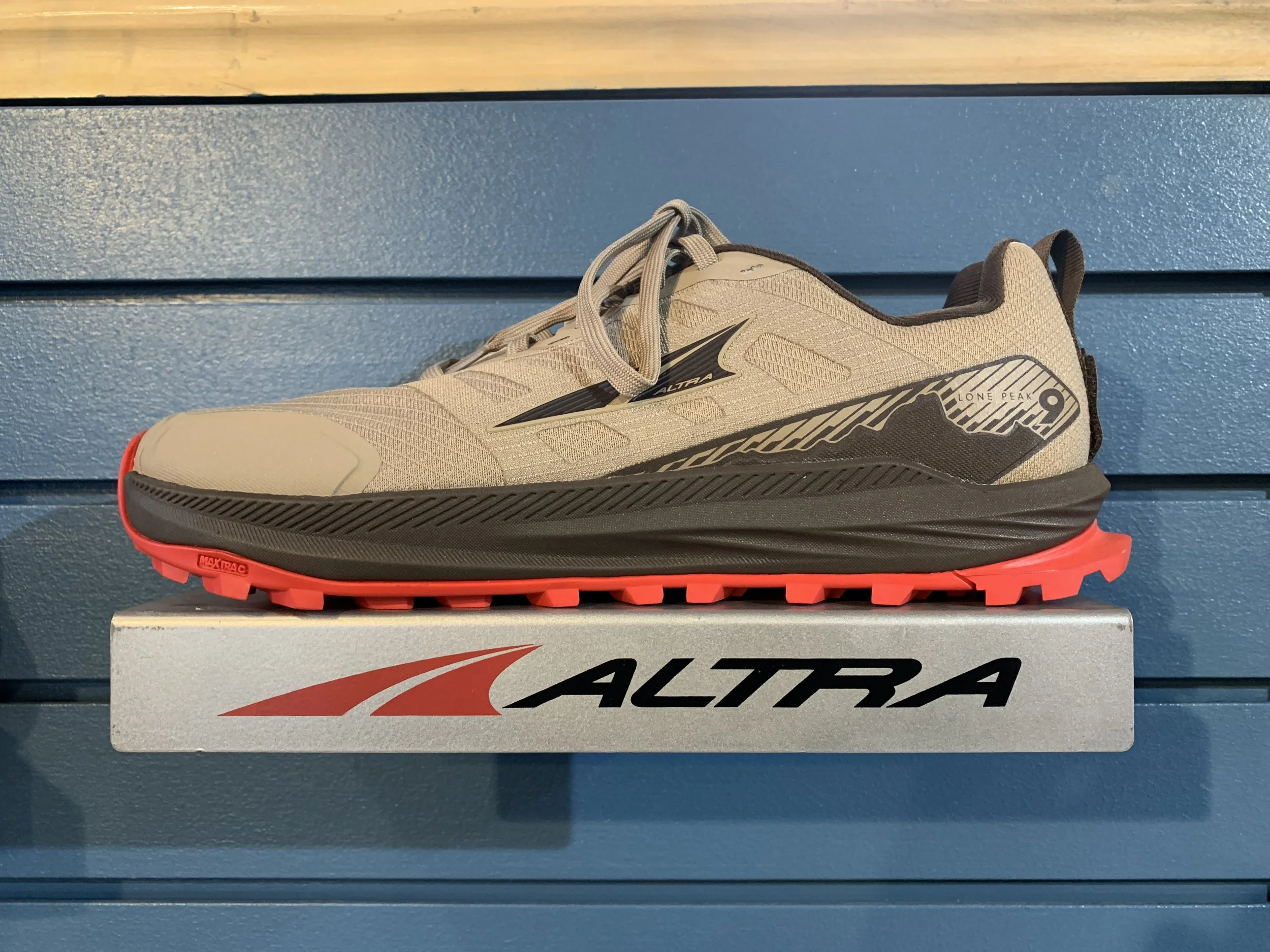 A beige and black Altra running shoe with red outsole, displayed on a silver box with Altra logo, against a blue wooden background.