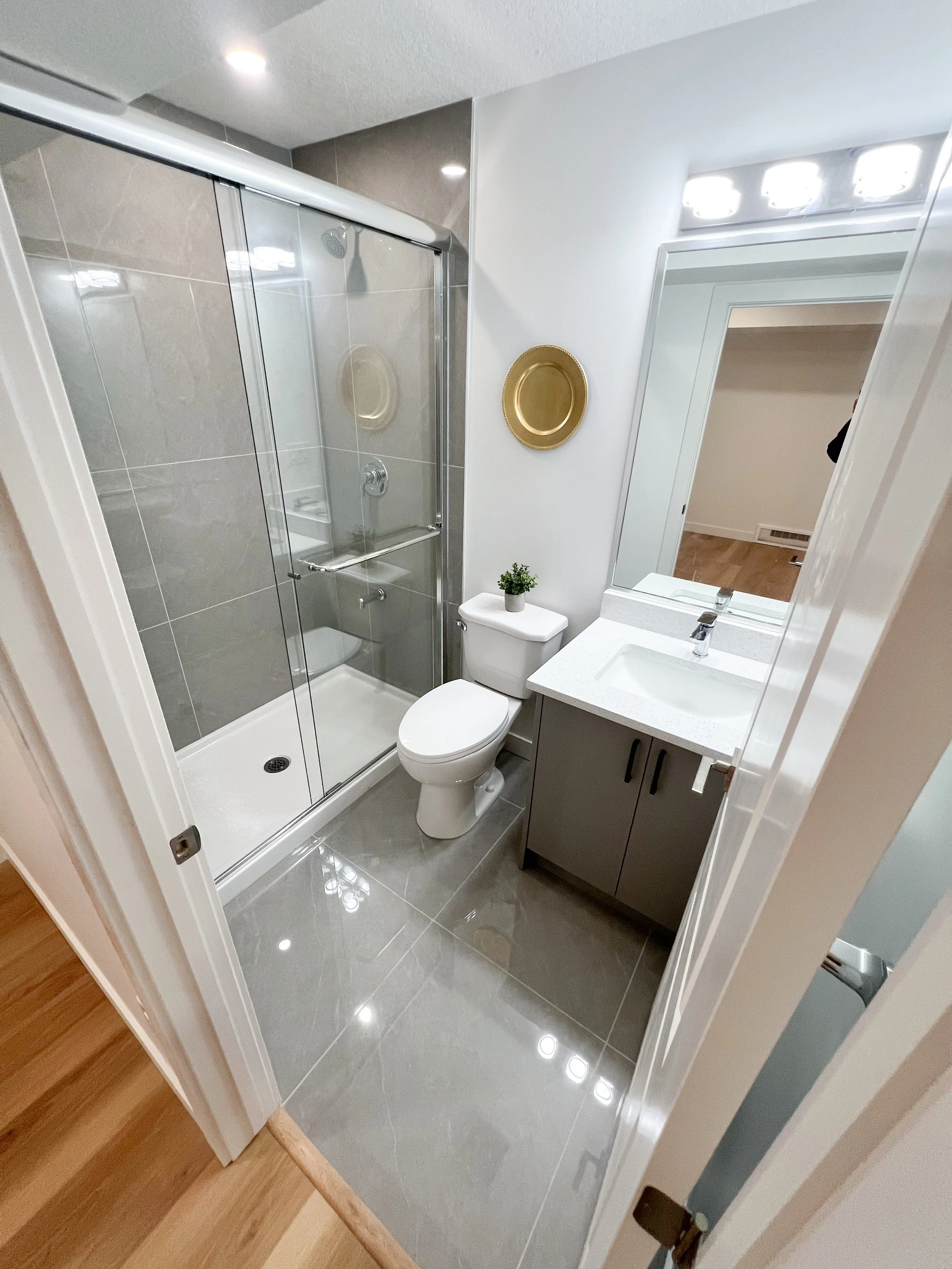 Small bathroom with a shower enclosure, a toilet, and a vanity with a sink. There is a large mirror above the vanity and decorative gold trays on the wall. The floors are tiled with gray tiles and there is a small potted plant on the toilet tank.
