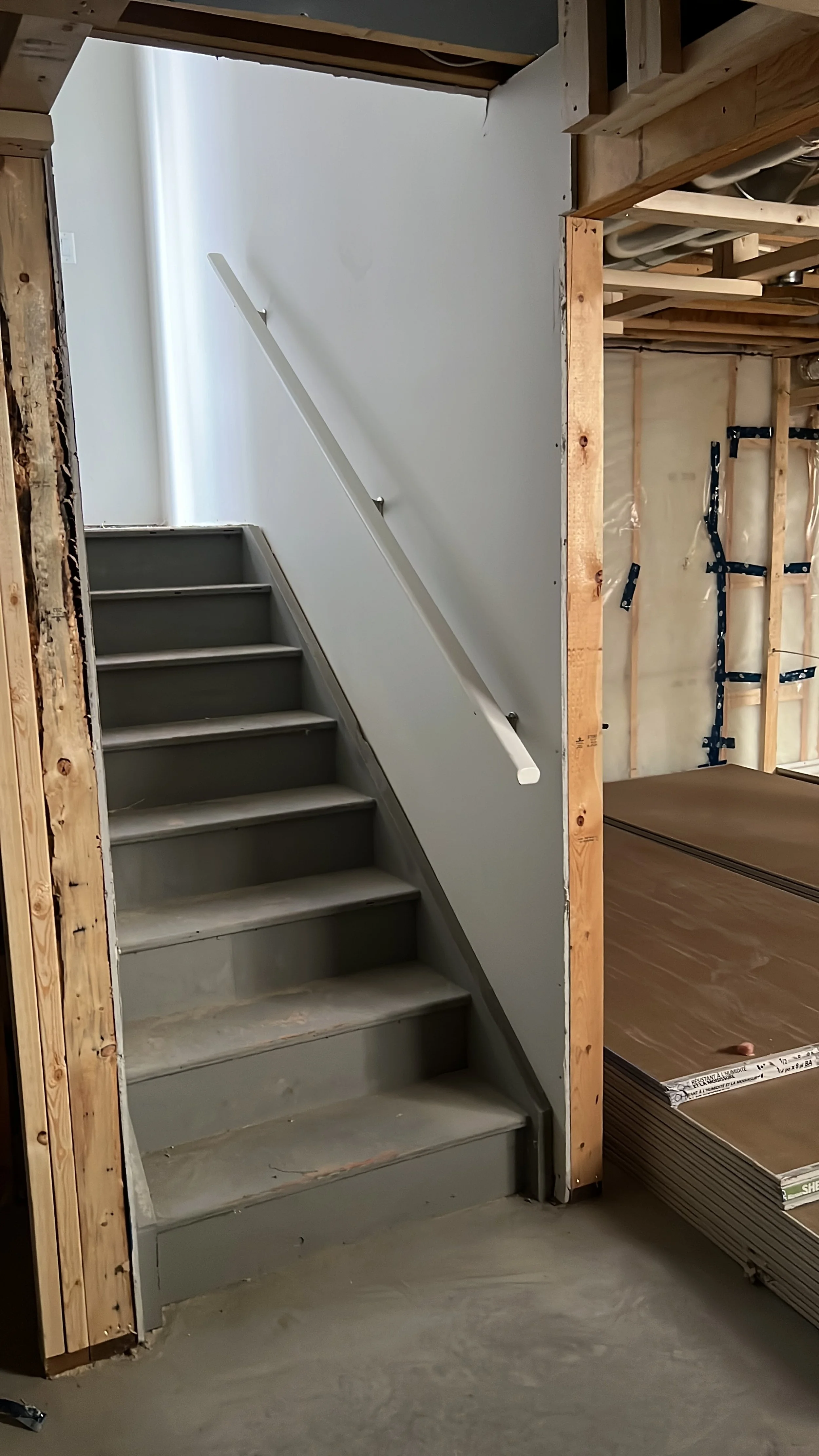 A partially finished interior space showing a staircase with gray steps, a white handrail, and surrounding framing with exposed wood and drywall work.
