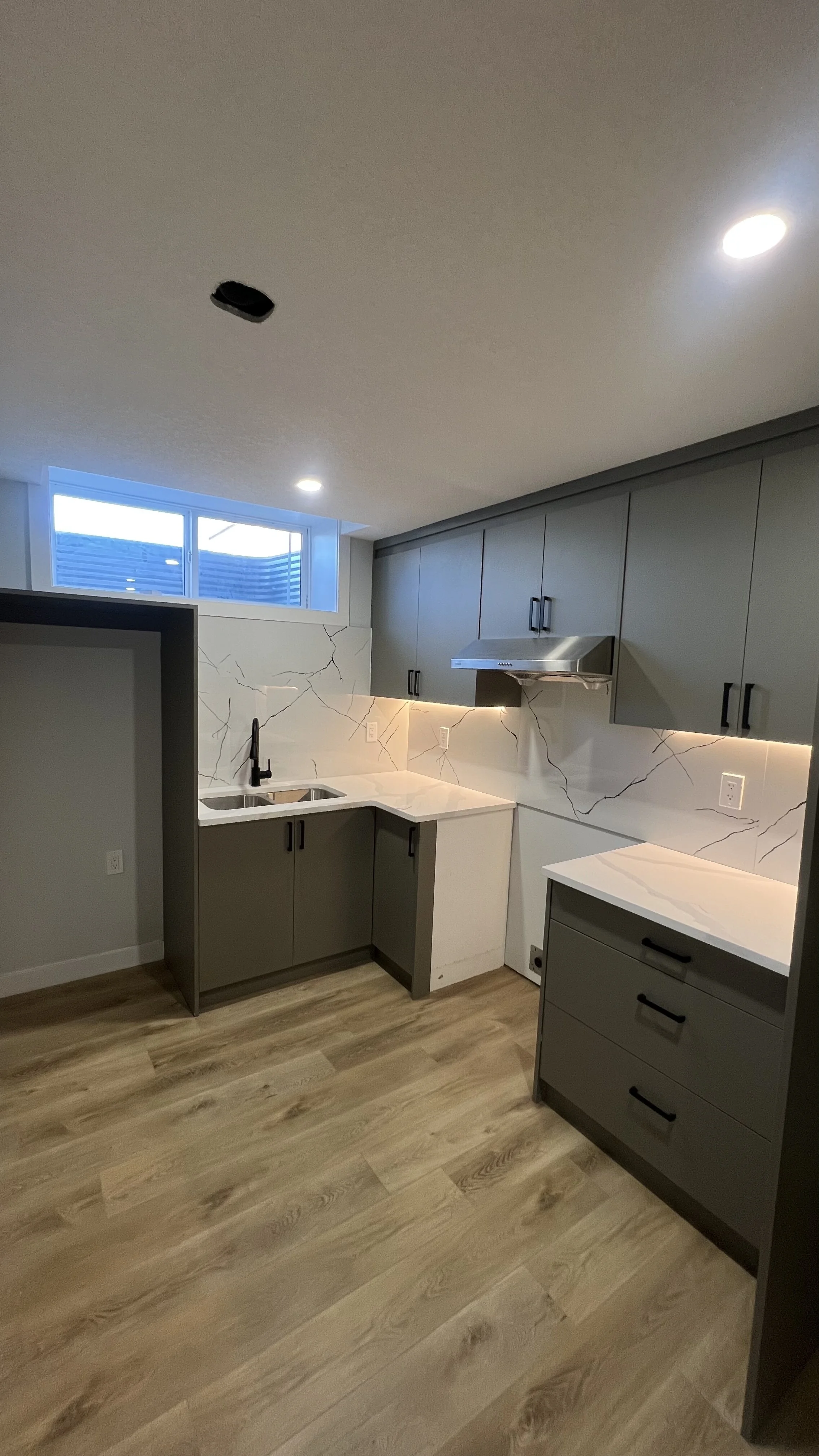 Modern kitchen with gray cabinets, white marble countertops, and a white marble backsplash with dark veining. It has a black faucet, wood flooring, and a window near the ceiling letting in natural light.