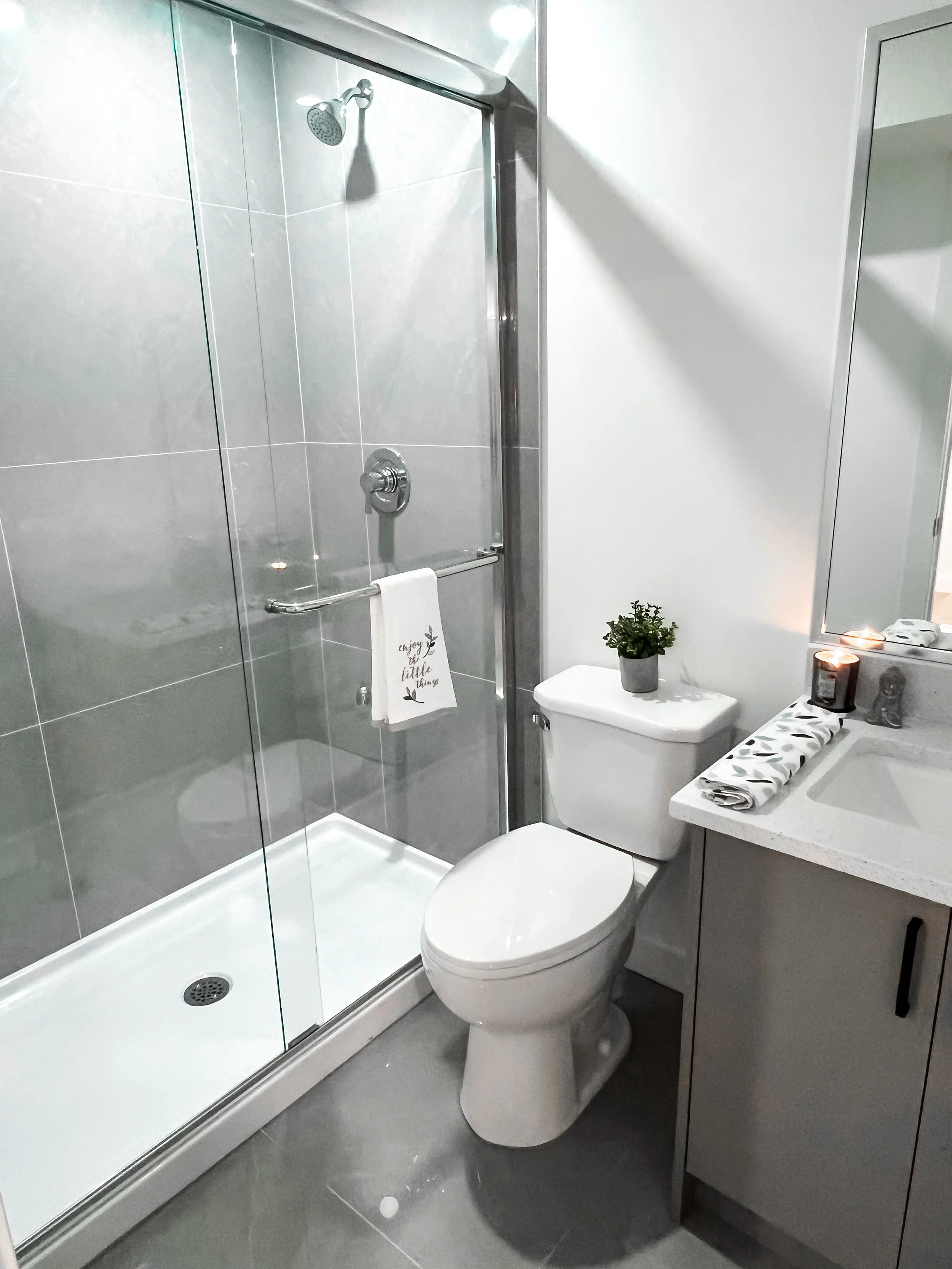 Modern bathroom with a glass shower enclosure, white toilet, gray cabinets, and a white sink with rolled towel and candles.