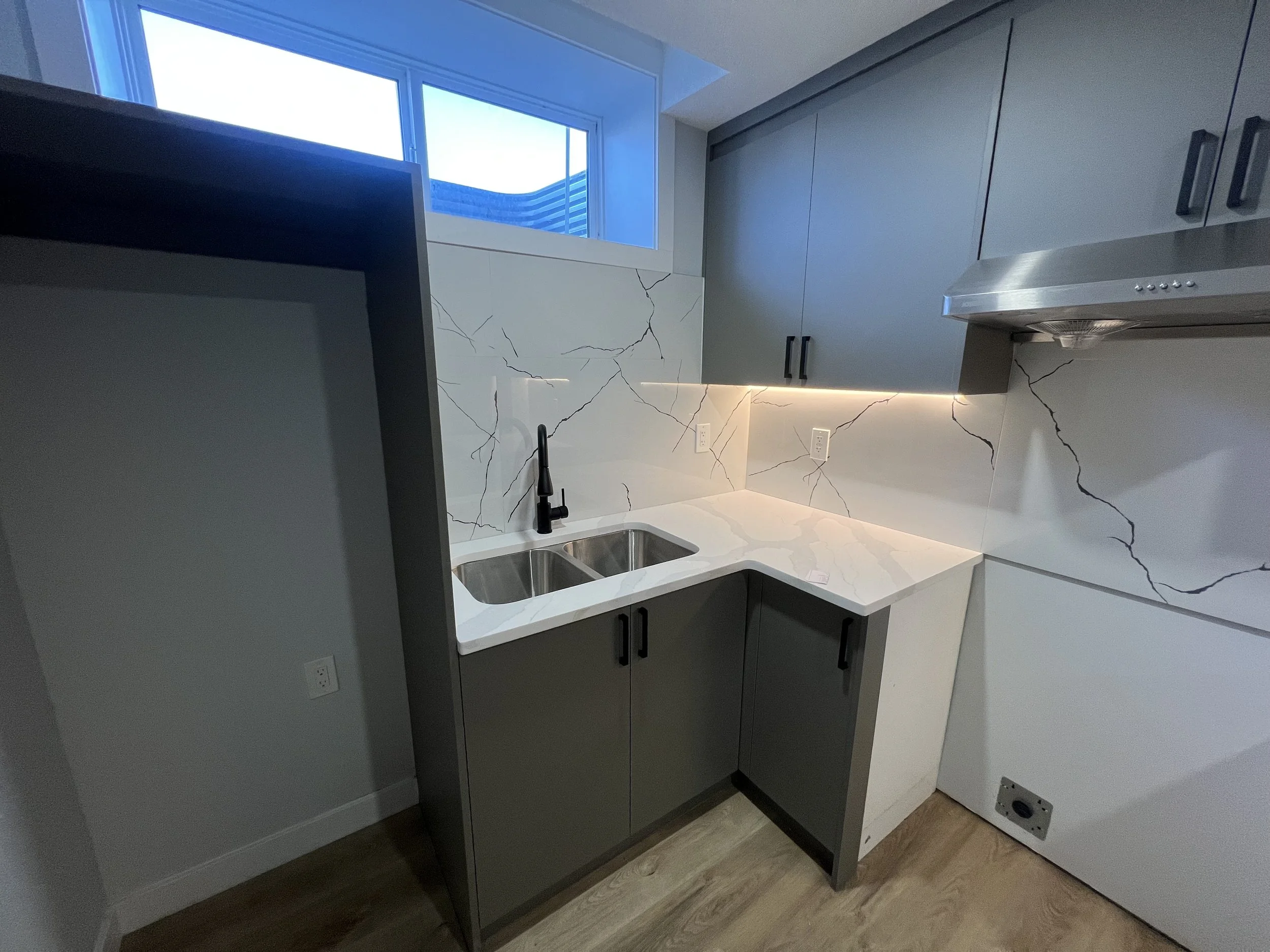 A modern kitchen corner with white marble countertops, gray cabinets, a double stainless steel sink, black faucet, and a large window overhead. The backsplash and part of the wall are covered with marble tiles, and there is under-cabinet lighting.