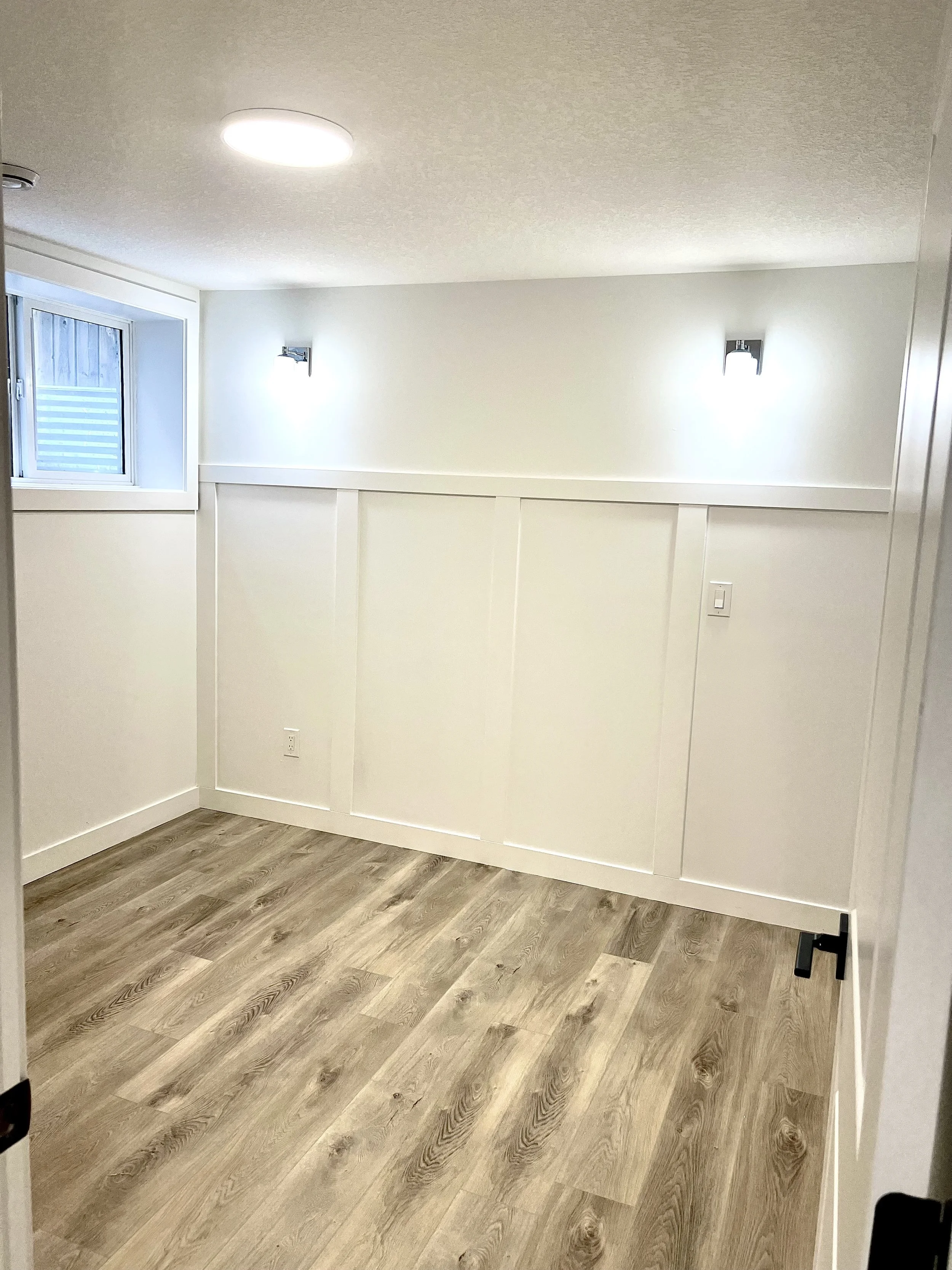 Empty room with wood flooring, white walls with paneling, a small window, and modern wall-mounted lights.