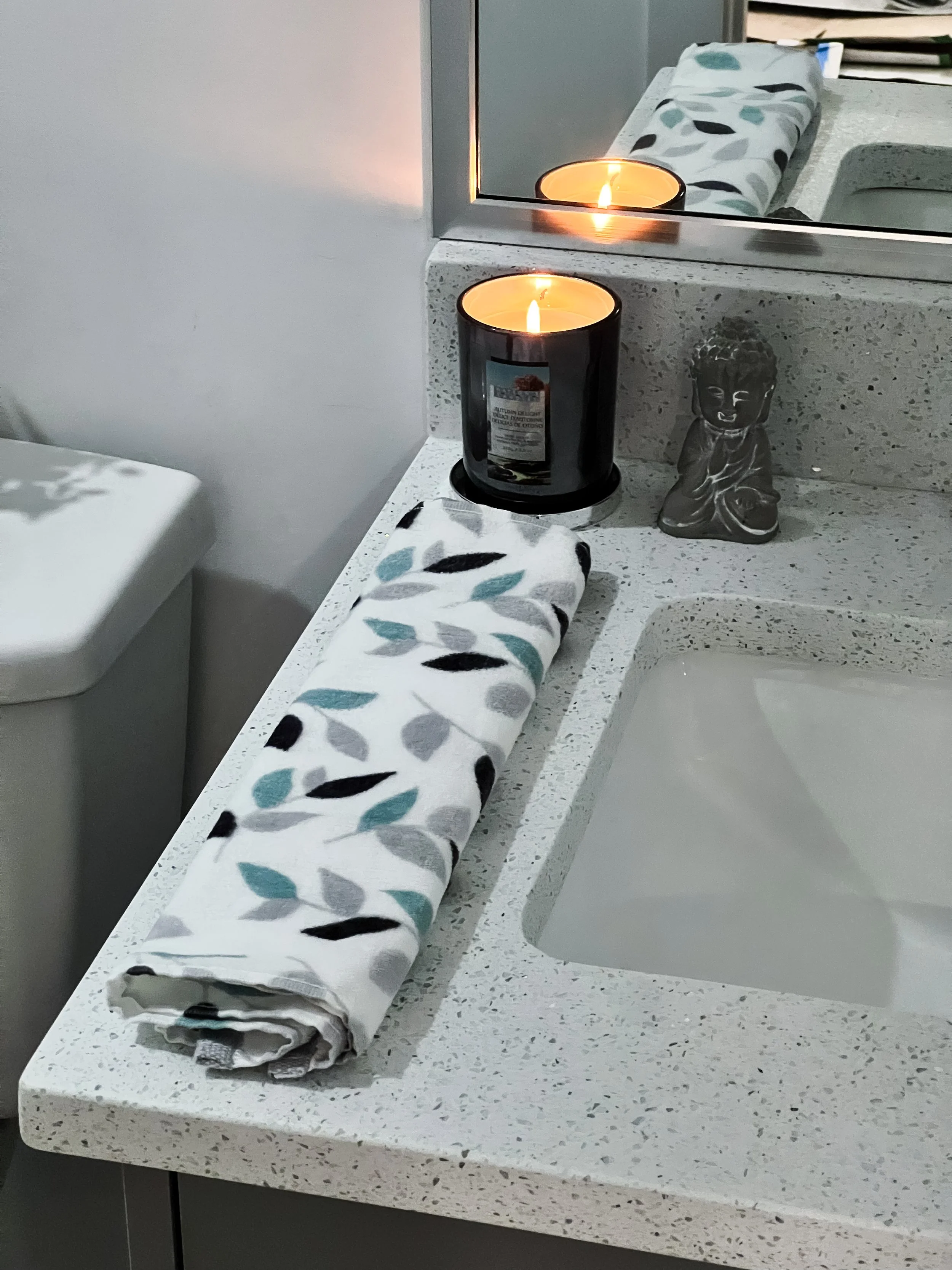 A rolled-up towel with a leaf pattern in black, gray, and teal colors on a speckled white bathroom countertop. Behind it, a lit candle in a black container and a small Buddha figurine are placed, with a mirror reflecting the candle and part of the co