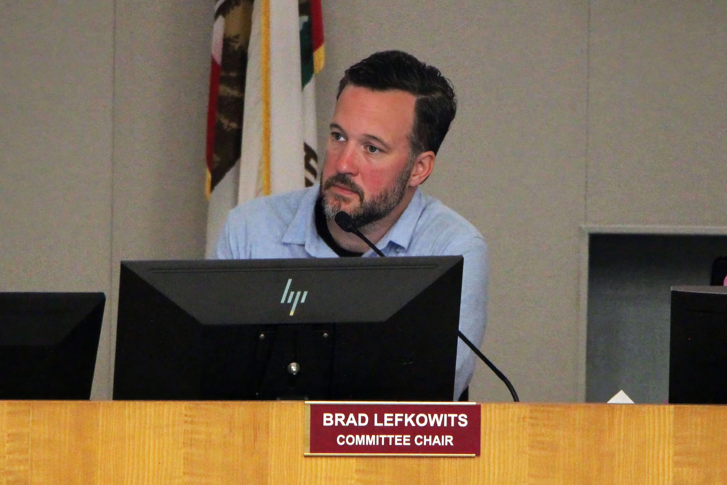 A man with dark hair and a beard sitting at a desk with multiple computer monitors. Behind him, there are flags, and a nameplate on the desk reads "Brad Lefkowitz, Committee Chair."