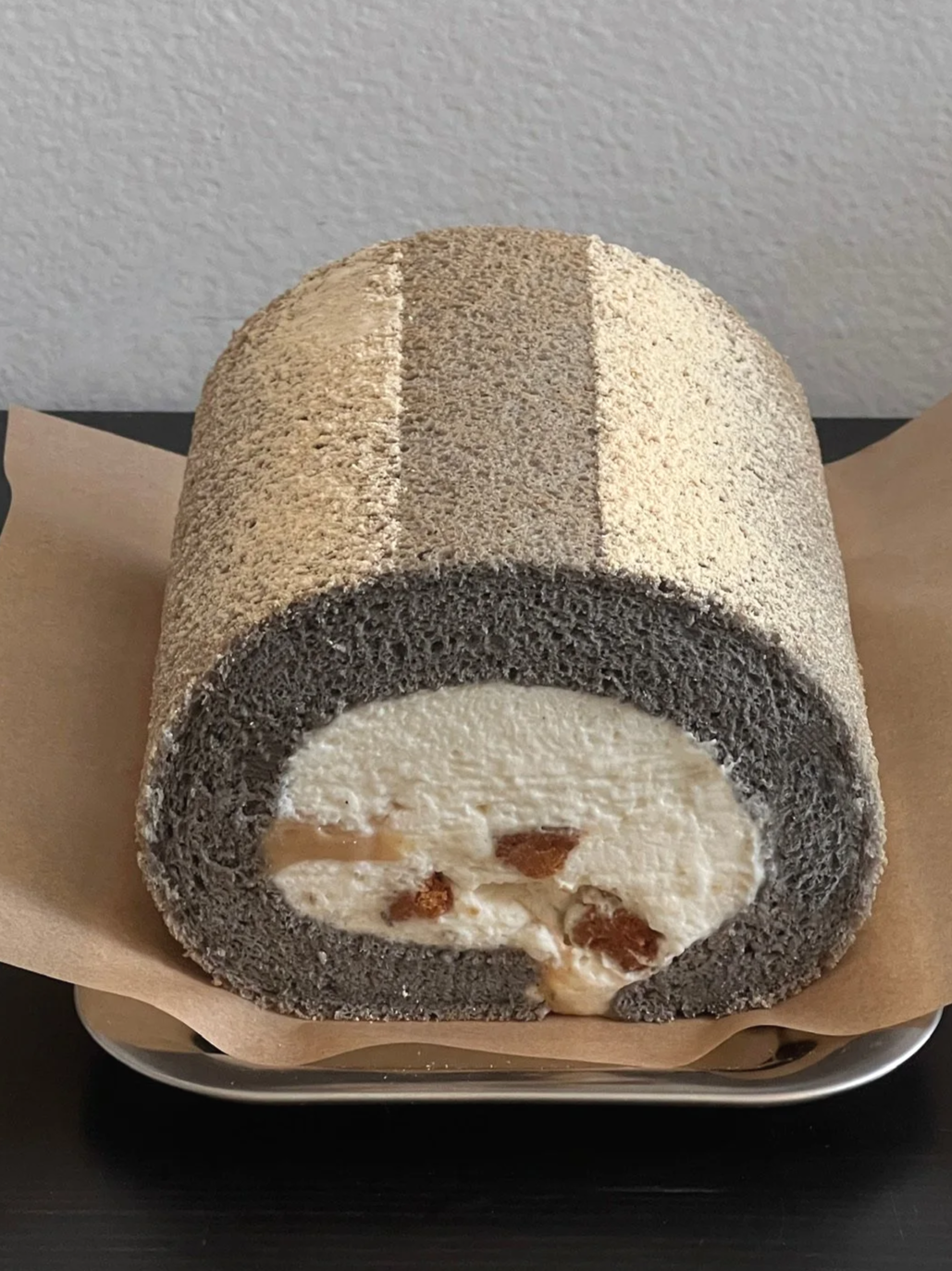 A black and white Swiss roll cake with a dark gray sponge, white creamy filling with raisins, sliced to show the interior, resting on a piece of parchment paper.