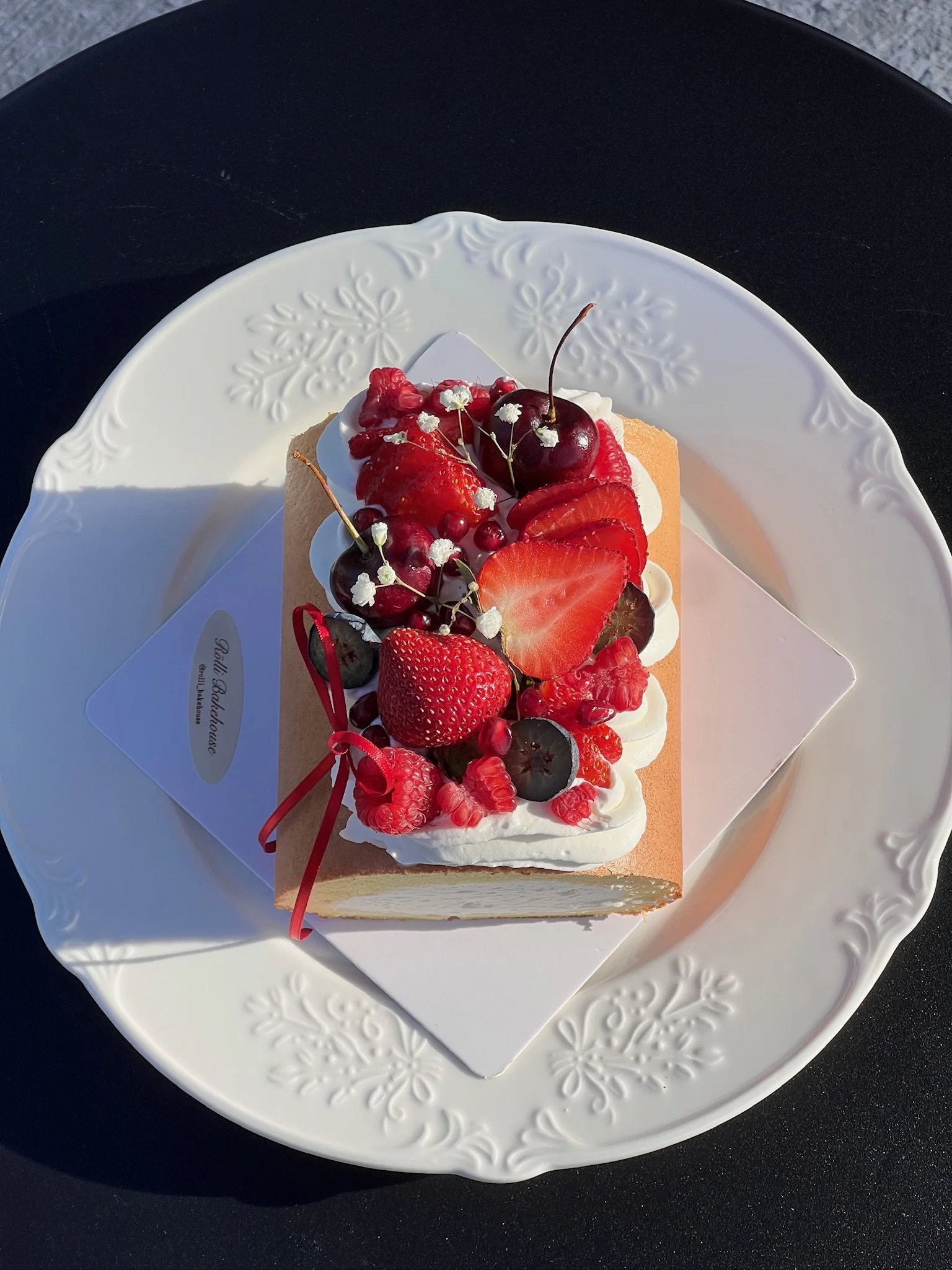 A cake decorated with fresh strawberries, cherries, blackberries, raspberries, and small white flowers on top. The cake has whipped cream frosting and is placed on a white ornate plate.