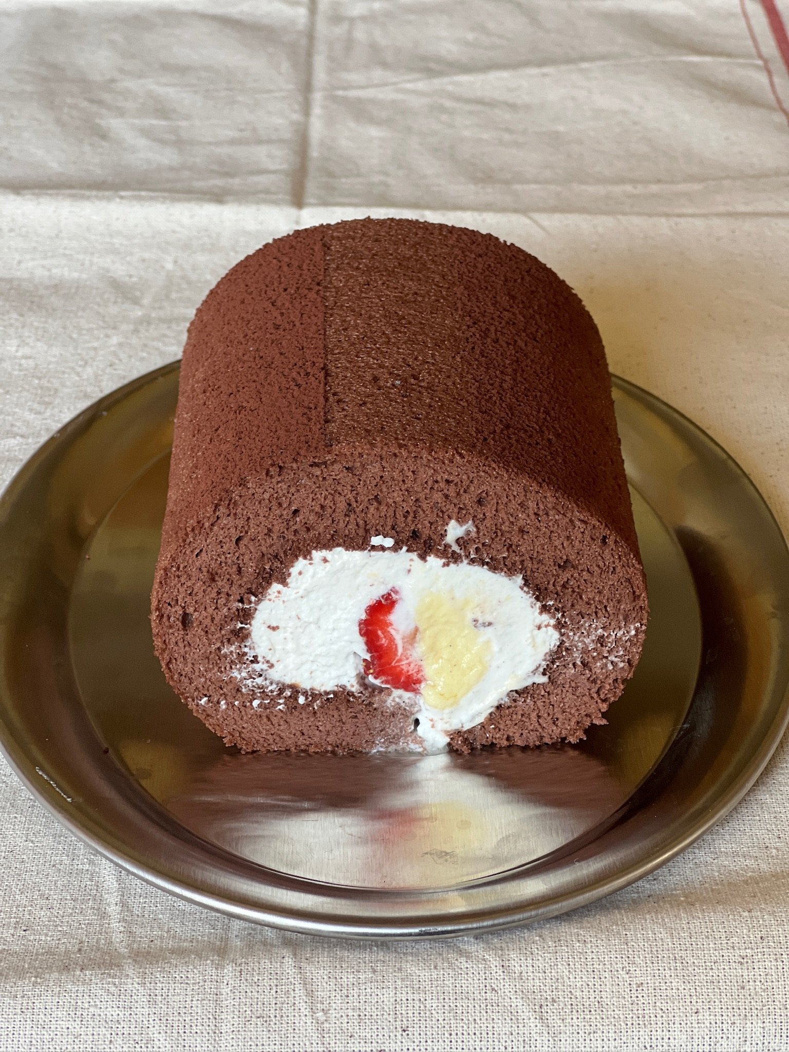 Chocolate Swiss roll cake with white cream filling and red and yellow fruit pieces, on a silver tray.