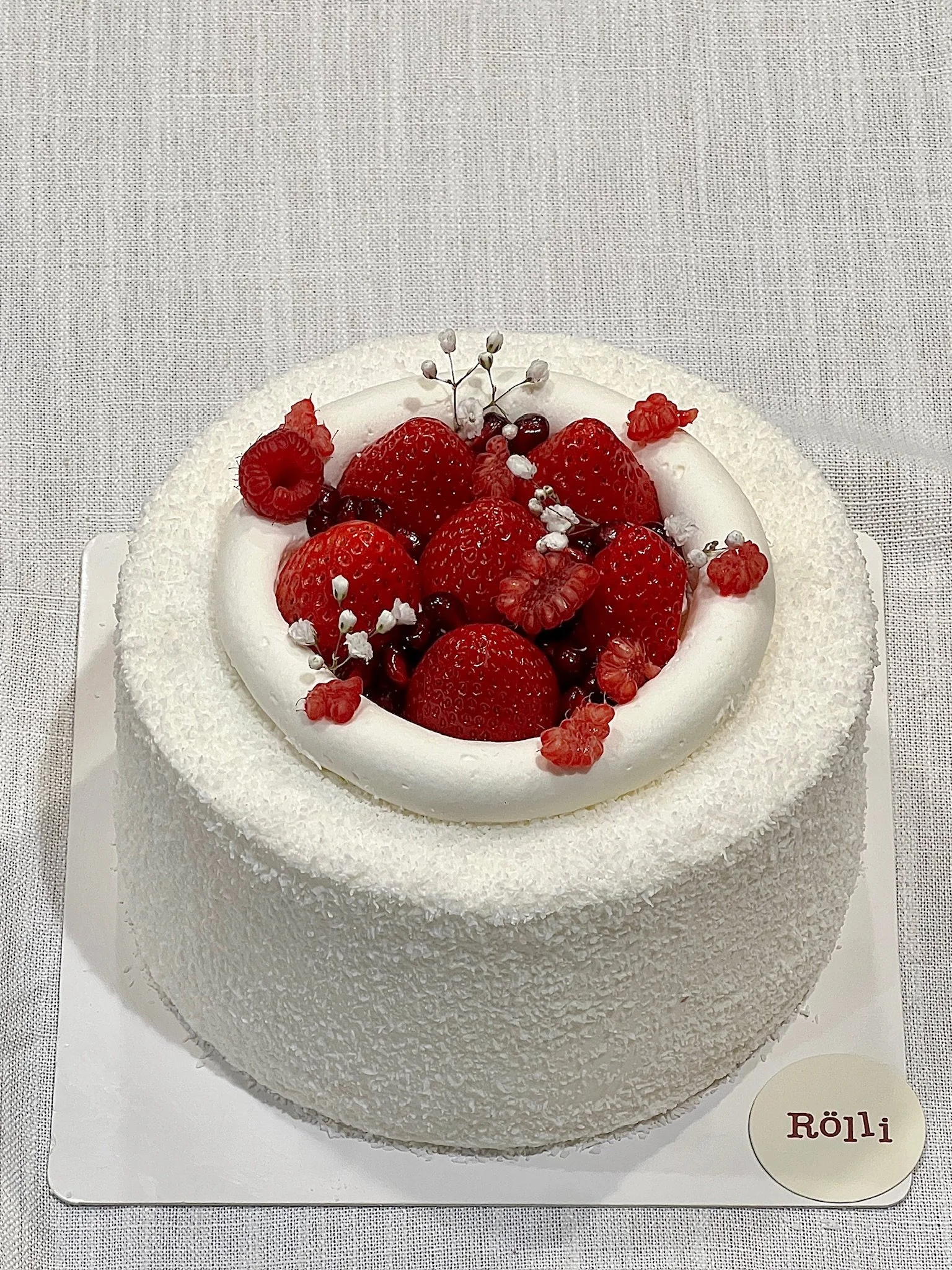 White cake with a coconut-flaked exterior, topped with fresh strawberries, raspberries, blackberries, and small white decorative flowers.