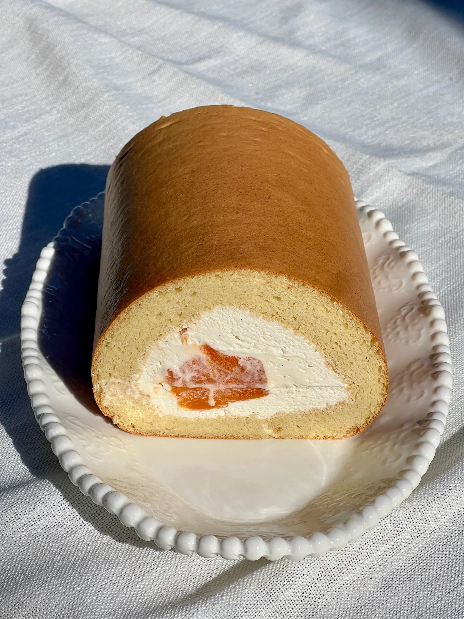 A slice of Swiss roll cake with cream and fruit filling on a decorative white plate, placed on a white tablecloth.