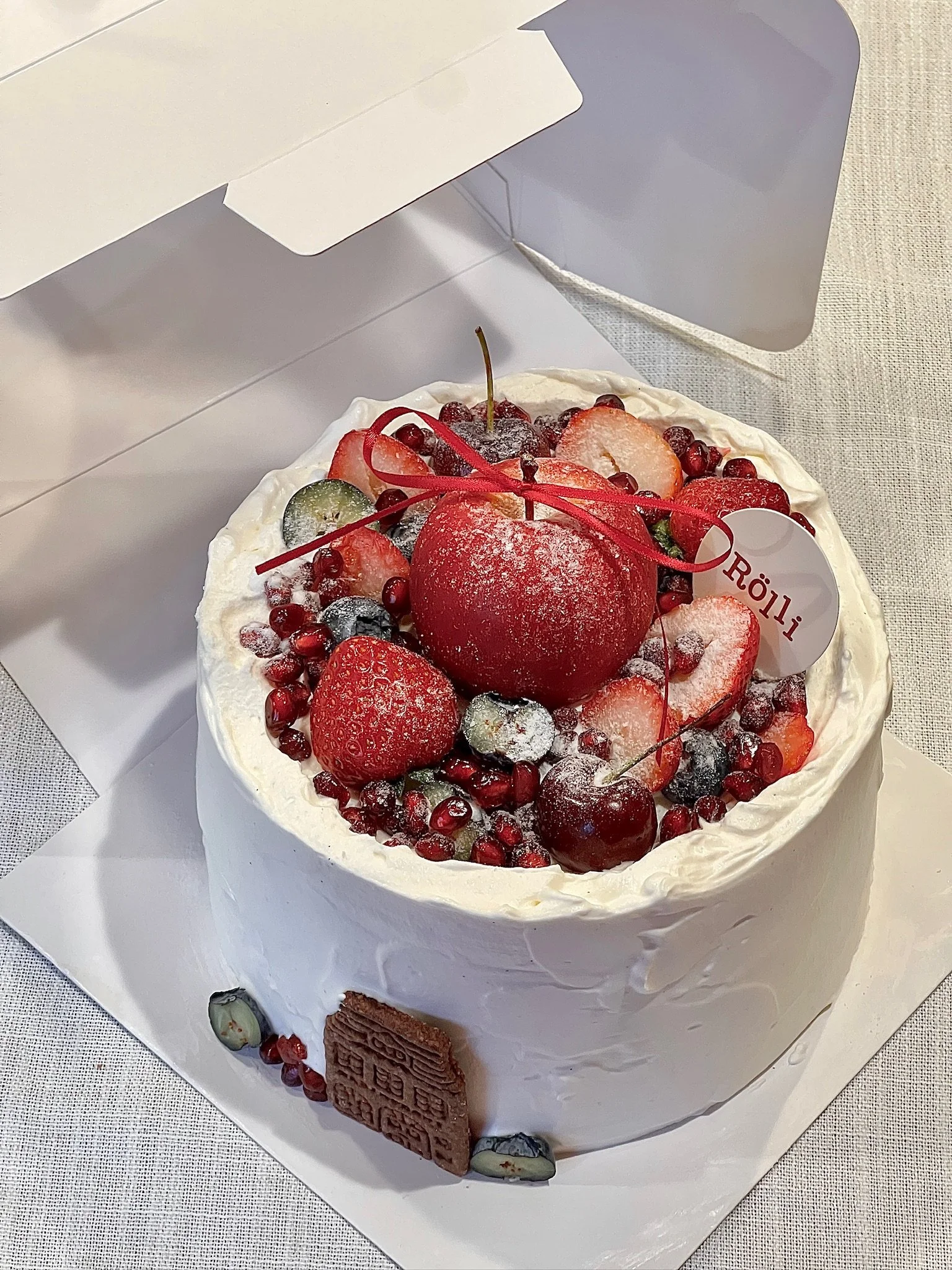 A round white frosted cake decorated with fresh strawberries, cherries, blueberries, pomegranate seeds, and a large red cherry on top. The cake is placed in an open white box on a light-colored table.