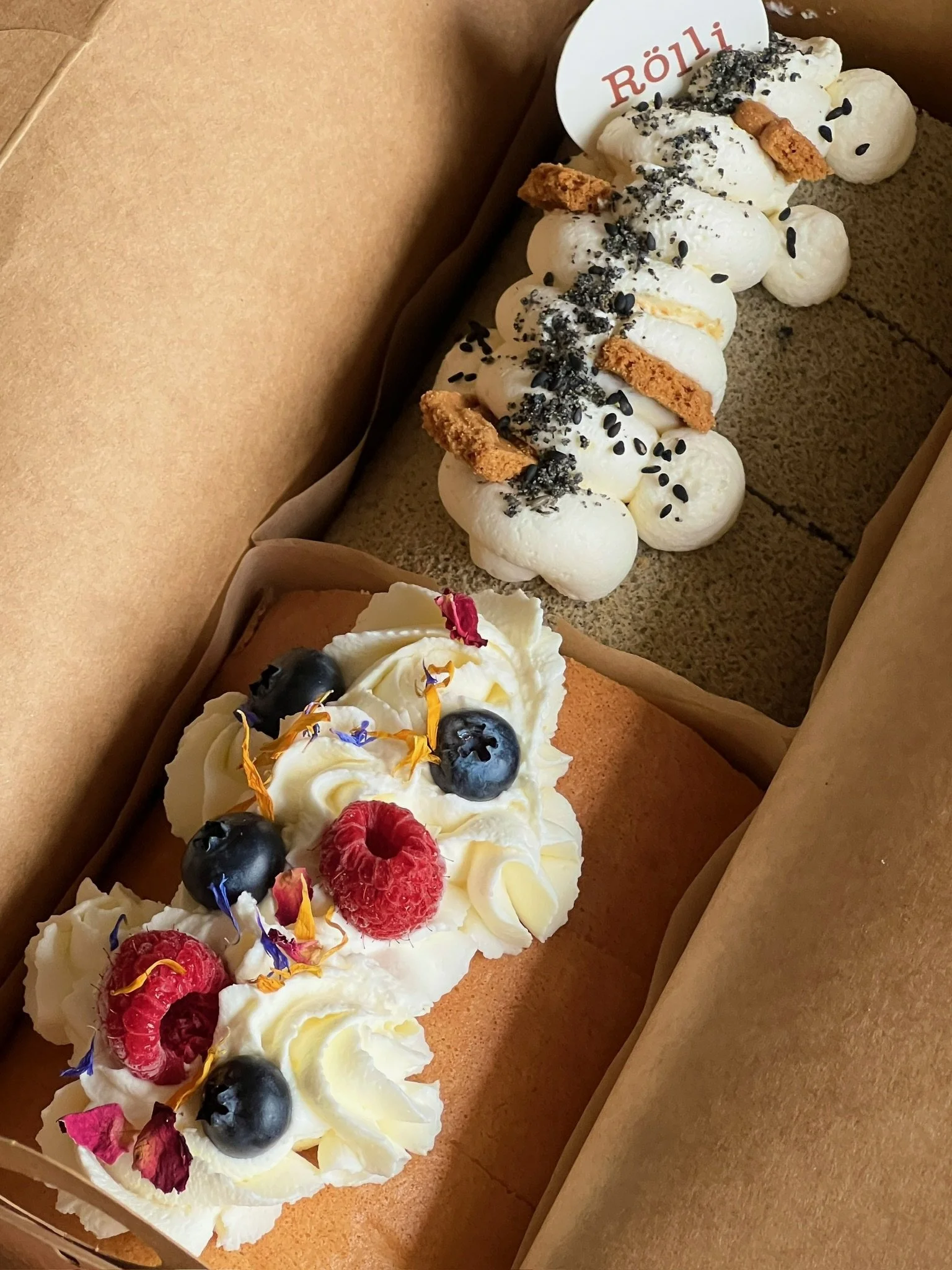 Two decorated cakes inside a cardboard box. The top cake has white frosting, cookie pieces, black sesame seeds, and black sprinkles. The bottom cake appears to be a cheesecake with whipped cream, blueberries, raspberries, and edible flower petals.