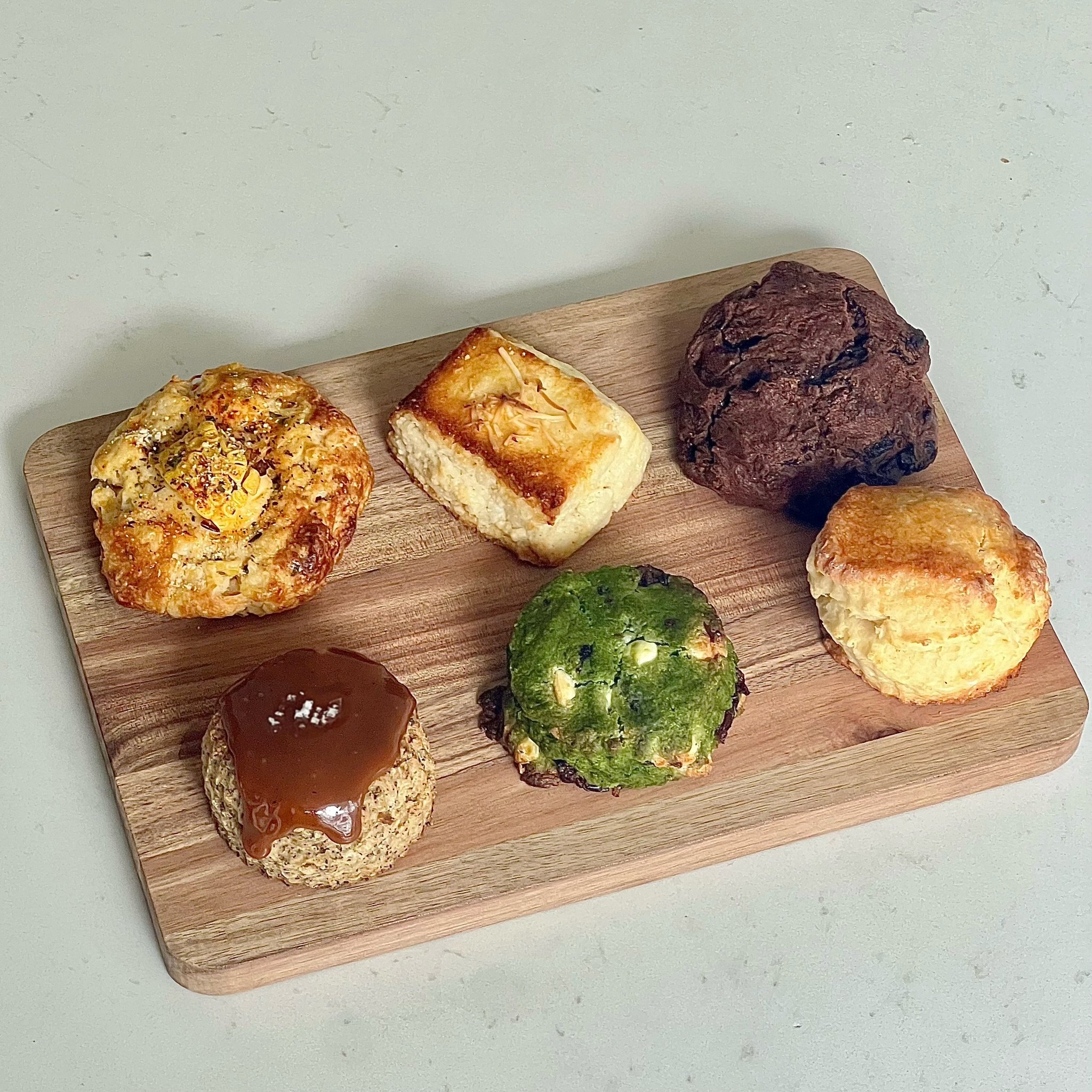 Assorted baked goods on a wooden tray, including muffins, a brownie, a cake square, a cookie with caramel, and a green muffin.