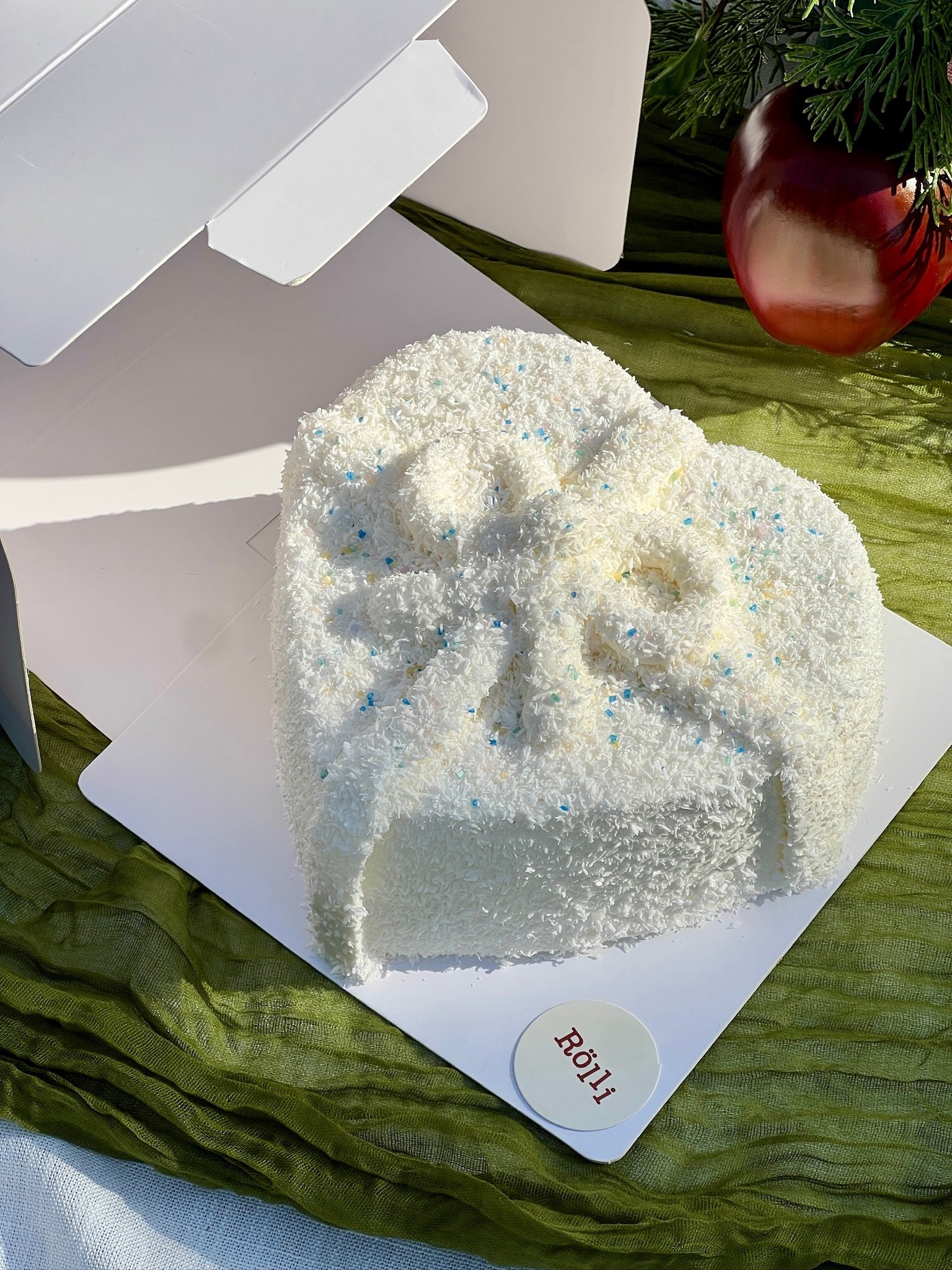 White heart-shaped cake decorated with shredded coconut and small blue and yellow sprinkles. The cake is in a white circle holder with the name tag 'Lili' on it, set on a white base with a green cloth underneath. There is a red apple and some greener