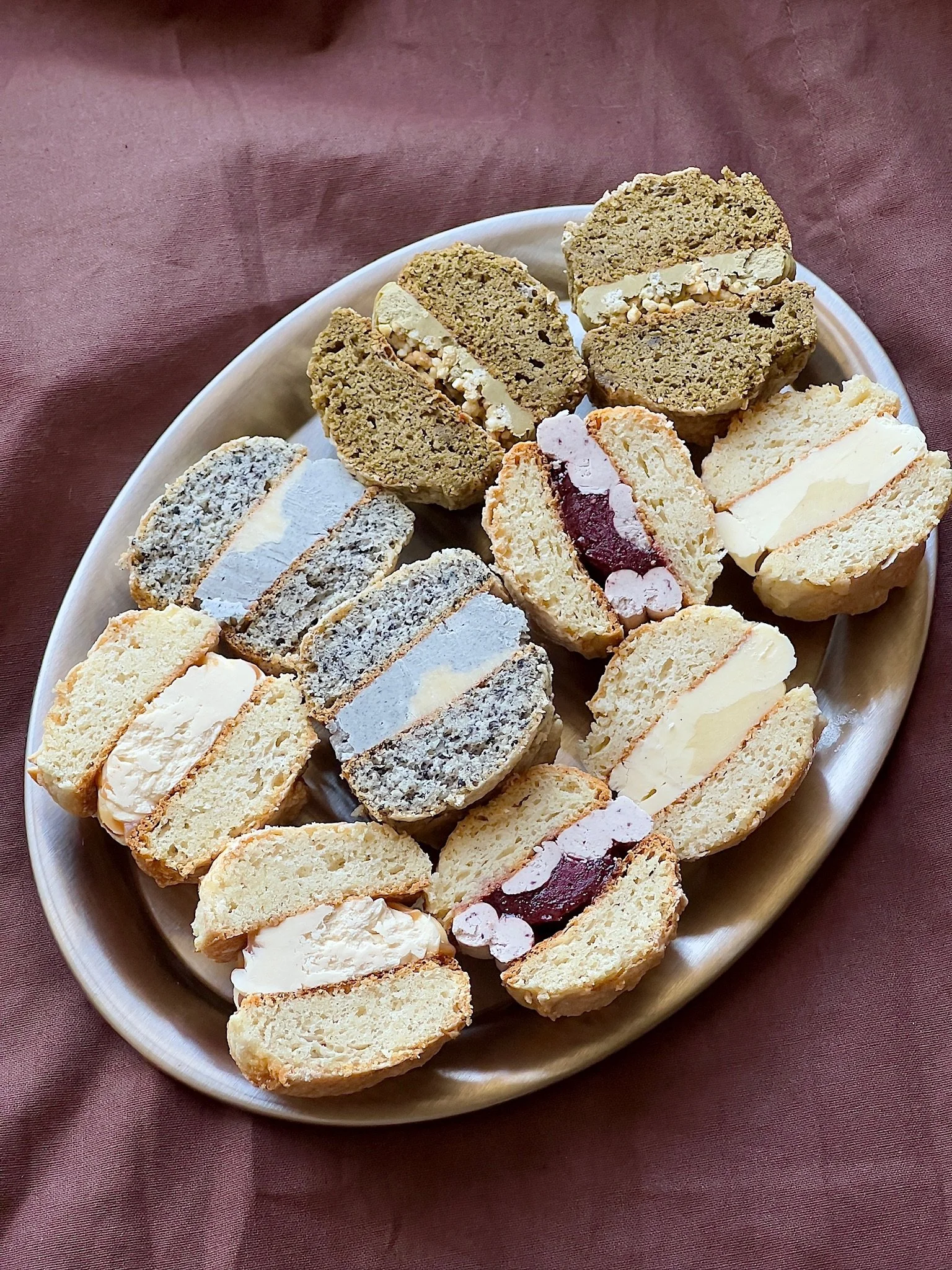 Assorted cookie sandwiches on a white oval platter, including varieties with cream, fruit filling, and cookie types, on a pink tablecloth.