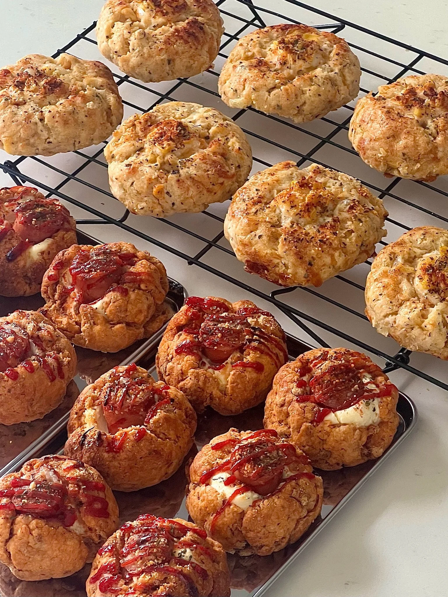 Assorted homemade baked goods including savory bread rolls on a cooling rack and sweet scones topped with ketchup on a tray.