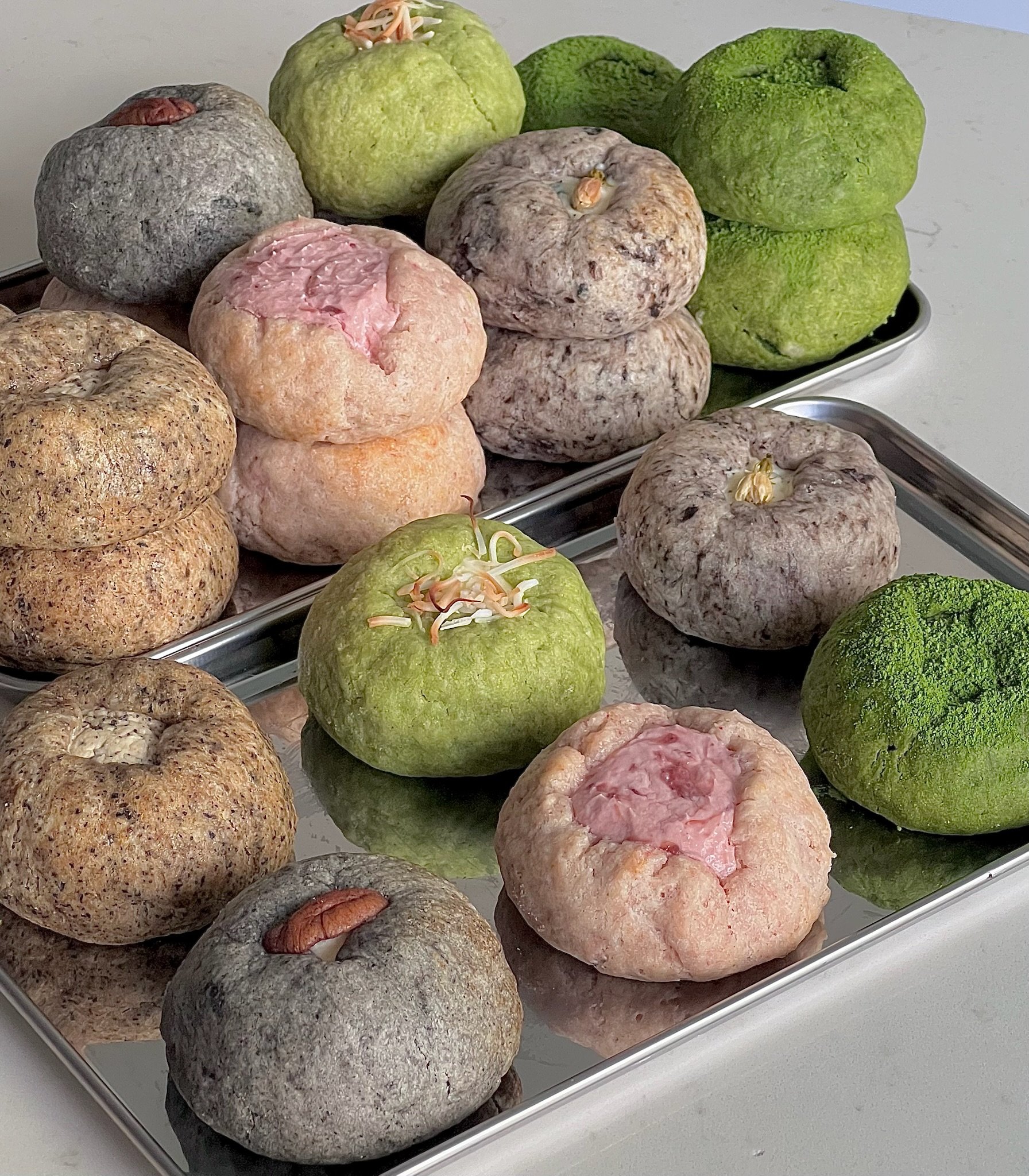 Variety of mochi ice cream balls on metal trays, some topped with sprouted beans or nuts, in green, pink, gray, beige, and brown colors.