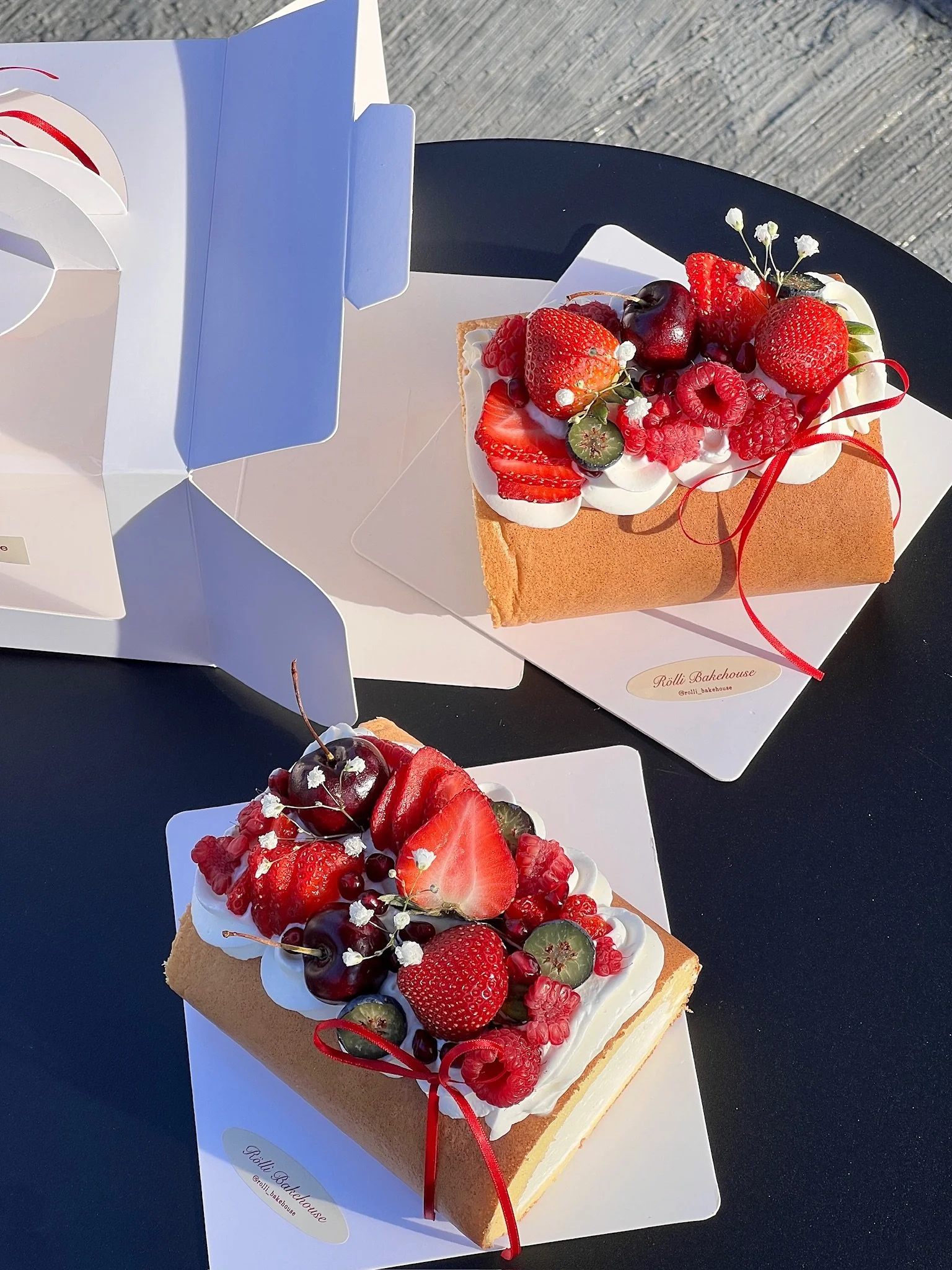 Two rectangular sponge cakes topped with whipped cream and fresh berries, including strawberries, cherries, raspberries, blueberries, and slices of kiwi, with decorative small white flowers and red ribbons, on white paper doilets on black tables.