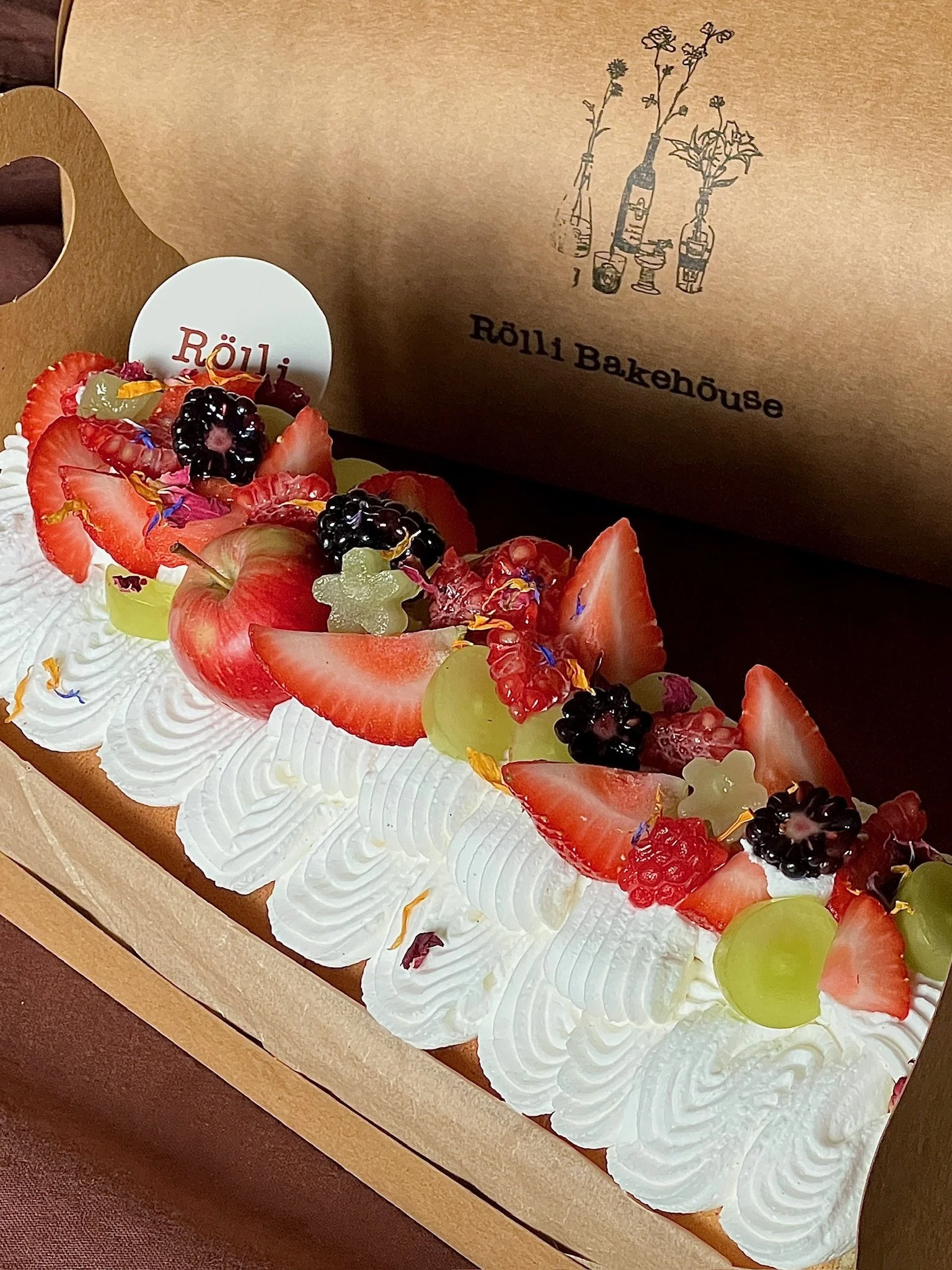 A fruit-topped cake with whipped cream and assorted fresh berries and grapes inside a Rölli Bakery box.