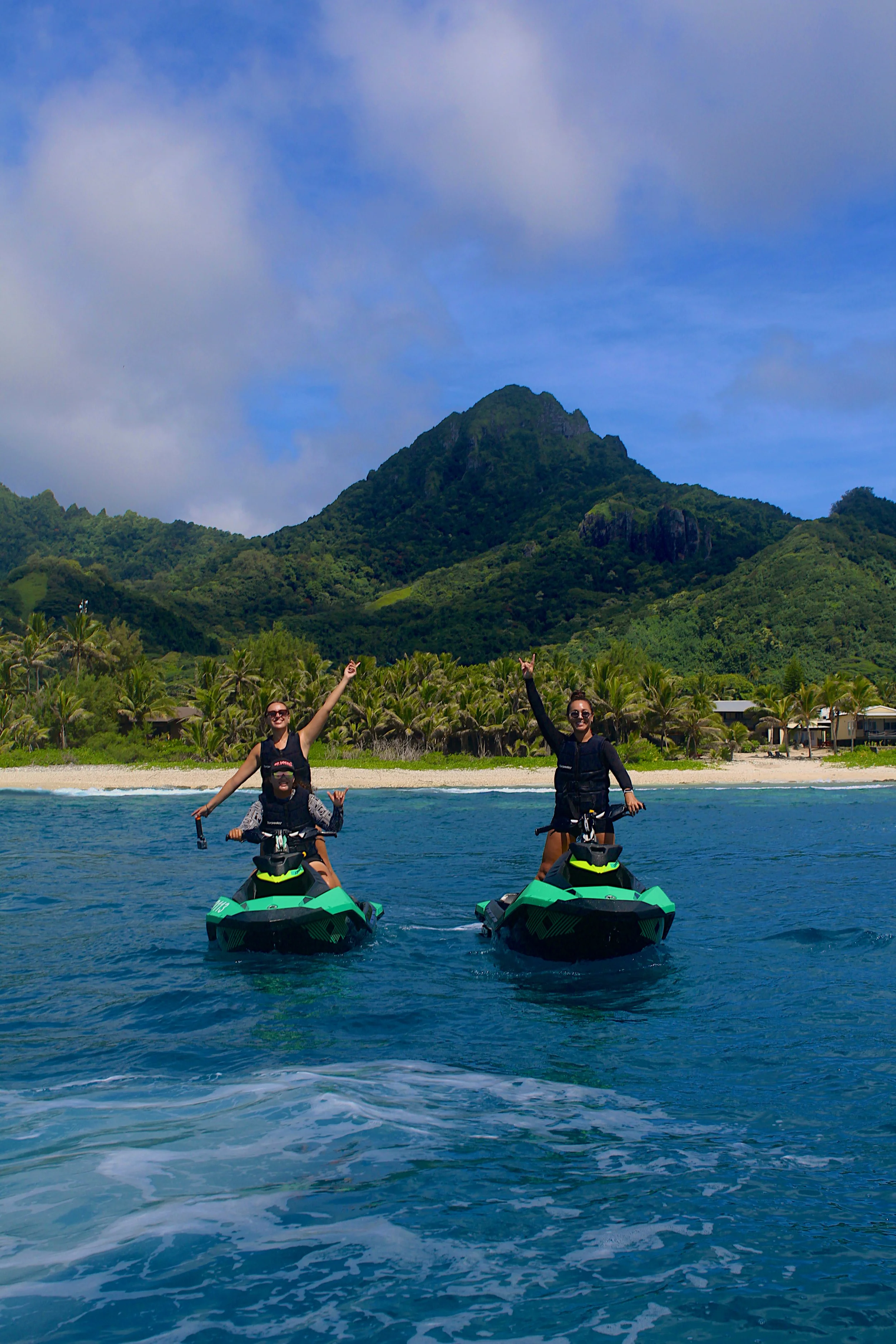 Two women riding jet skis on the water near a tropical beach with green mountains and trees in the background, both women are smiling and making peace signs.