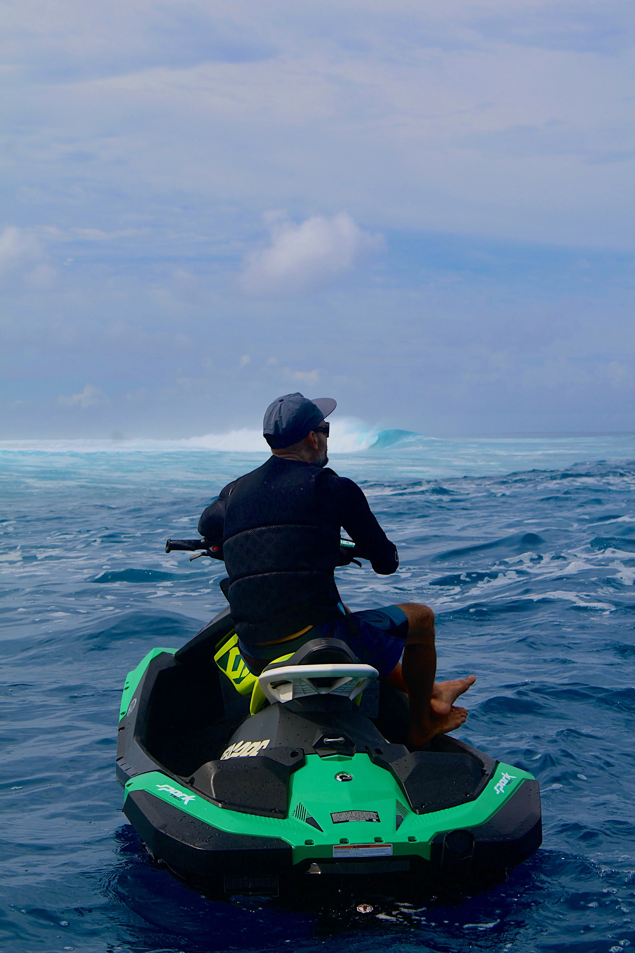 A man sits on a green and black jet ski on the ocean, facing the water, with a wave forming in the distance under a cloudy sky.