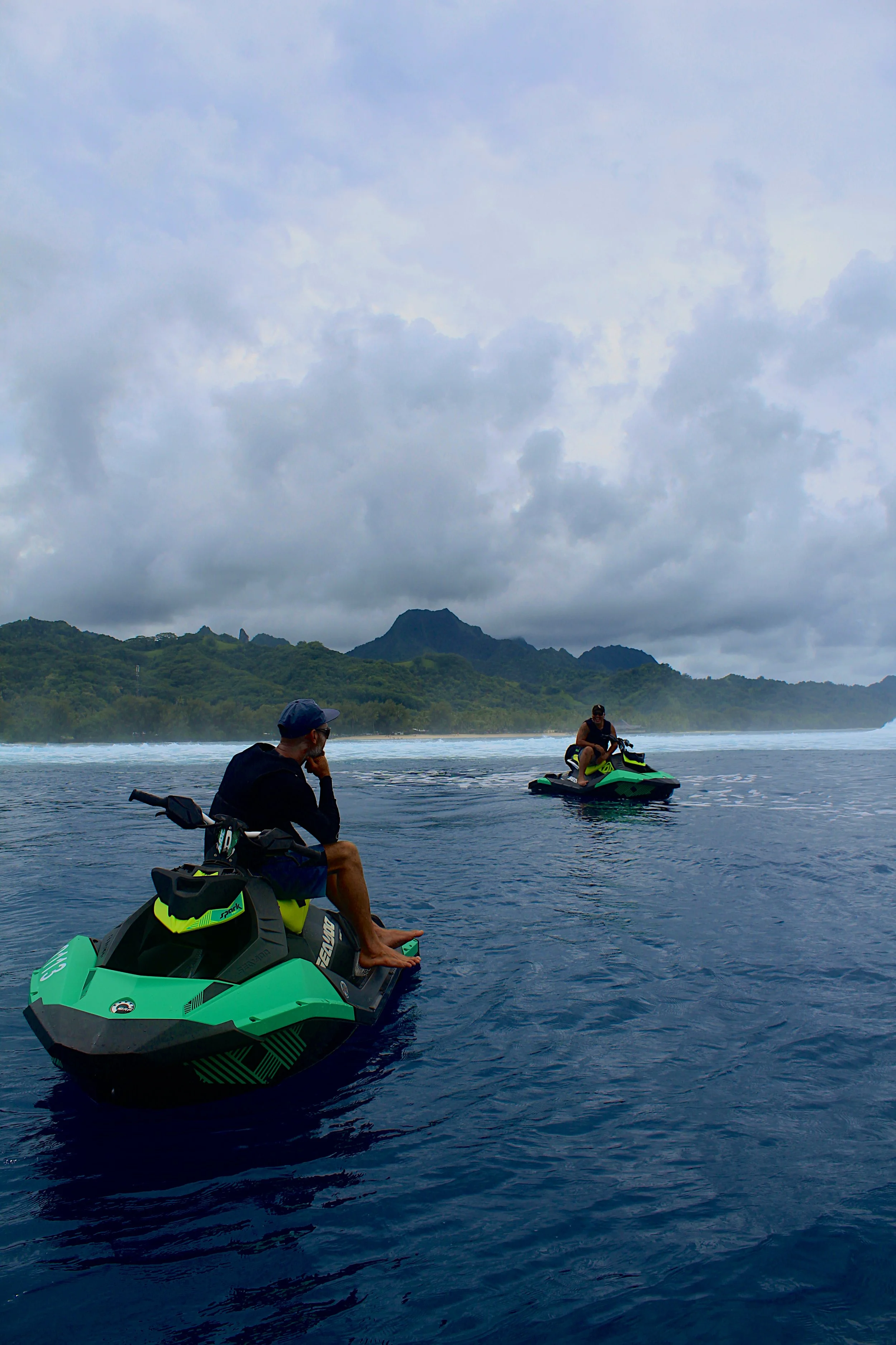 Two people riding jet skis on a calm ocean with a green mountainous landscape and cloudy sky in the background.