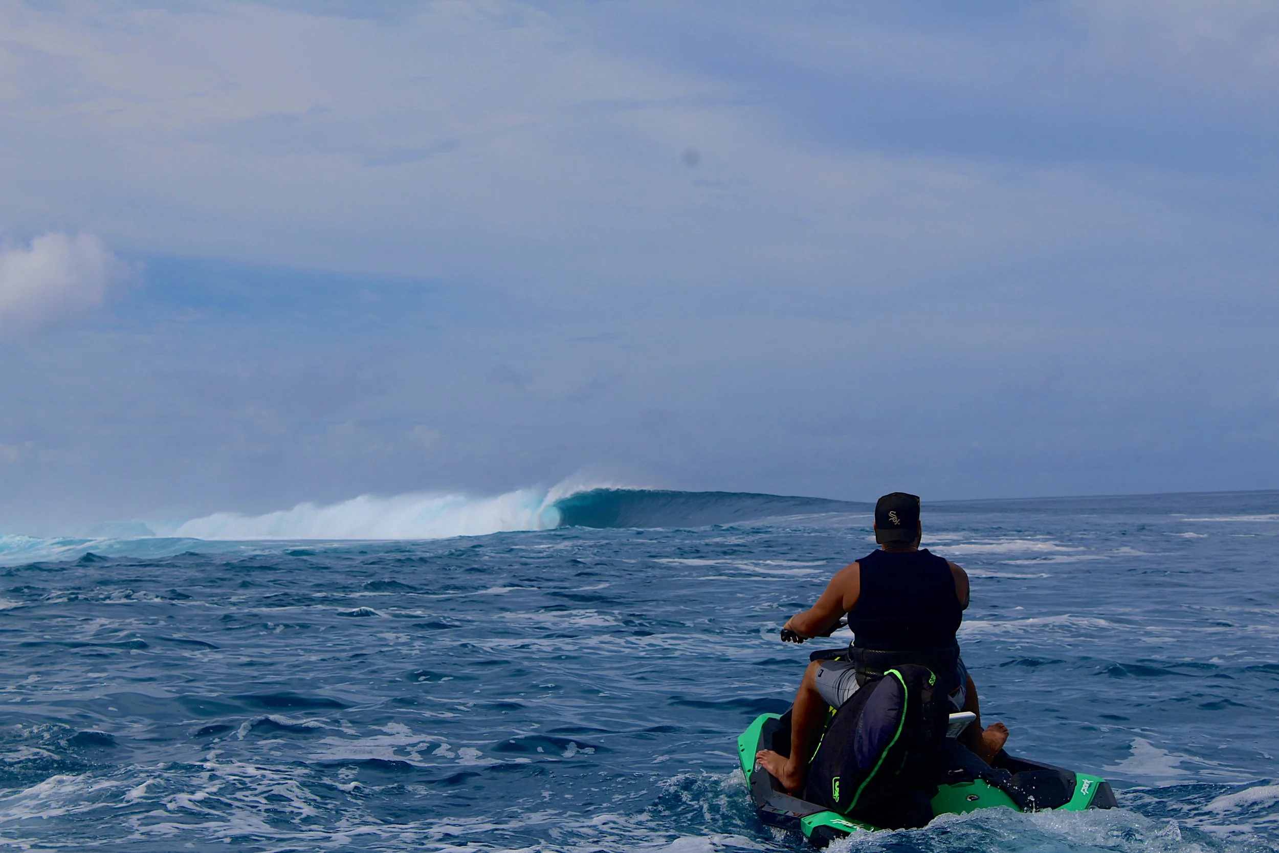 A person riding a jet ski on the ocean, facing a large wave in the distance, with an overcast sky.