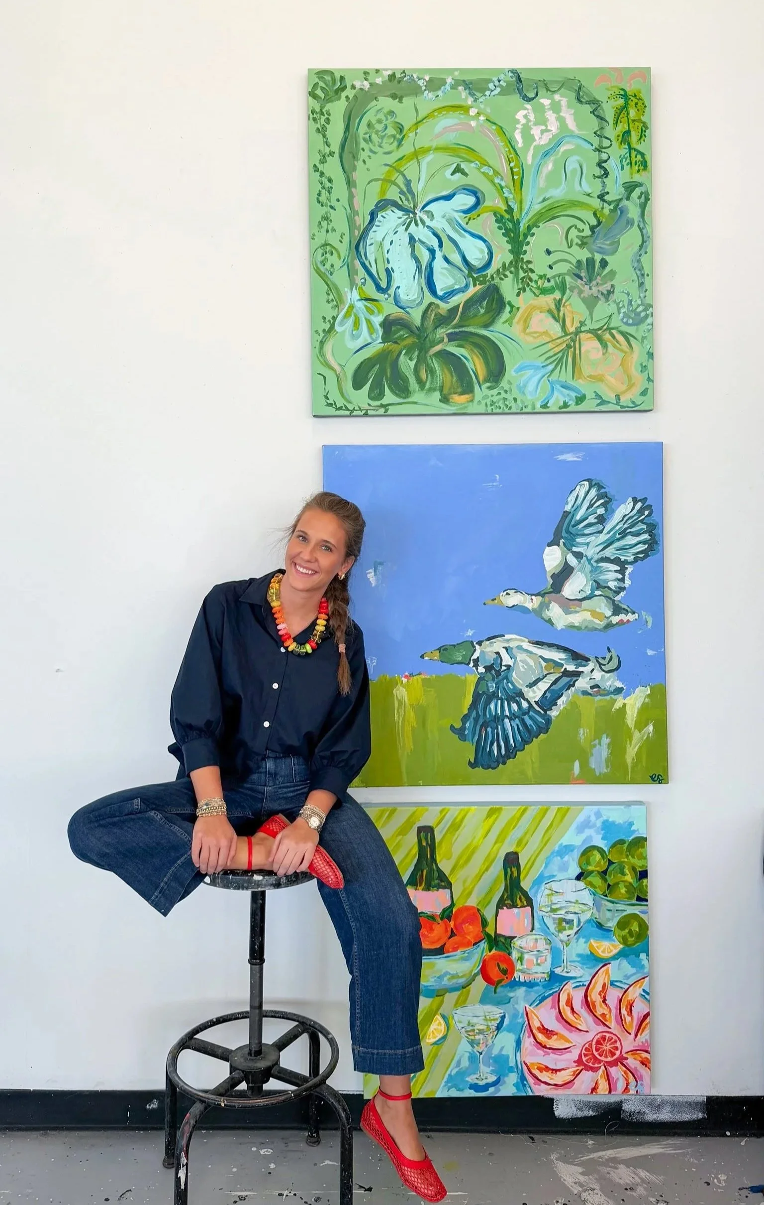 A woman with braided hair sitting on a black stool with her left leg crossed over her right, wearing red high heels, dark jeans, a navy blue shirt, and a colorful beaded necklace, at an art gallery with four abstract paintings of birds and fruits on a white wall behind her.