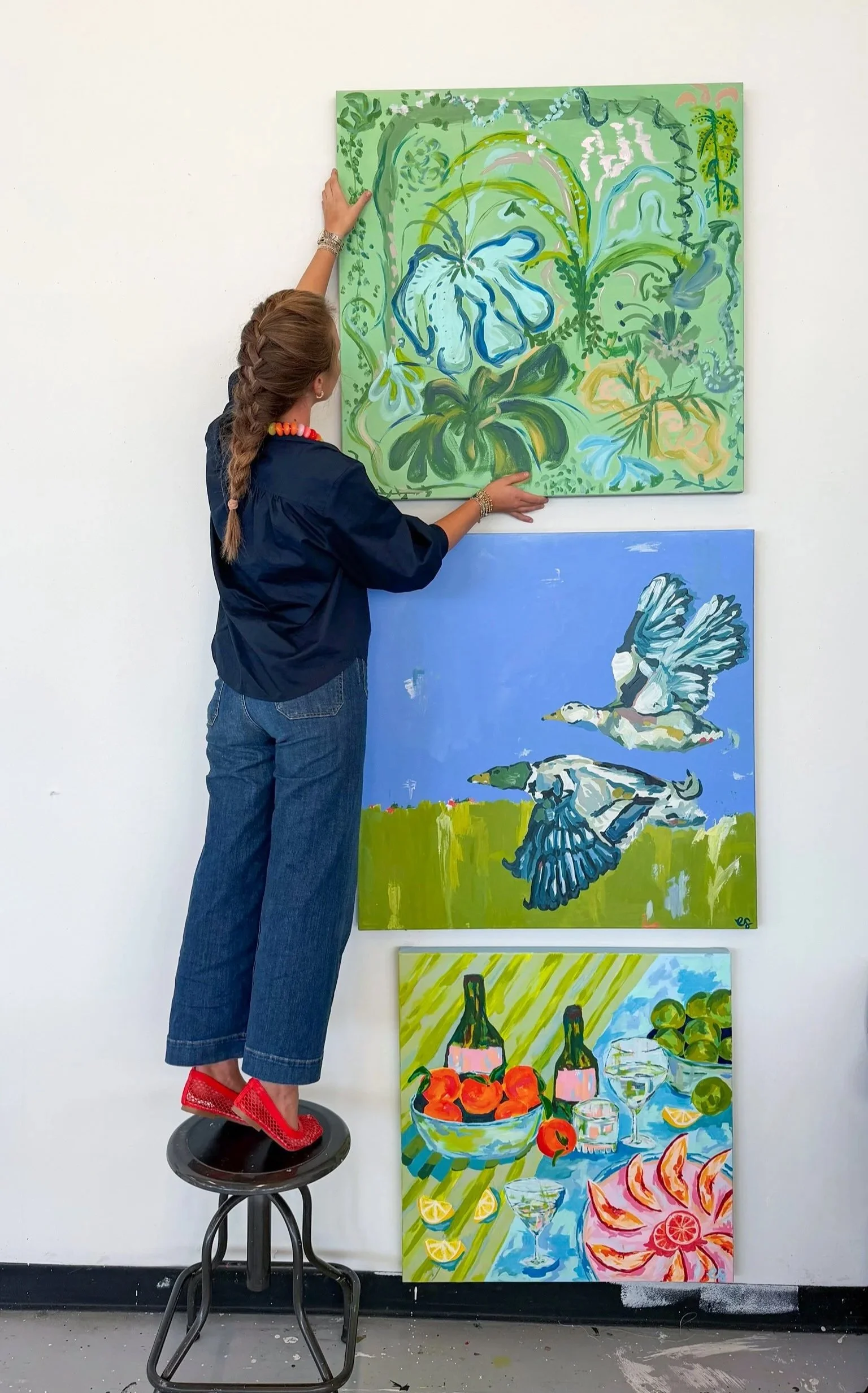 An art gallery with a woman standing on a stool to reach a large colorful painting of green foliage and flowers. Below are additional paintings of birds flying over a landscape and a still life of fruits and drinks.