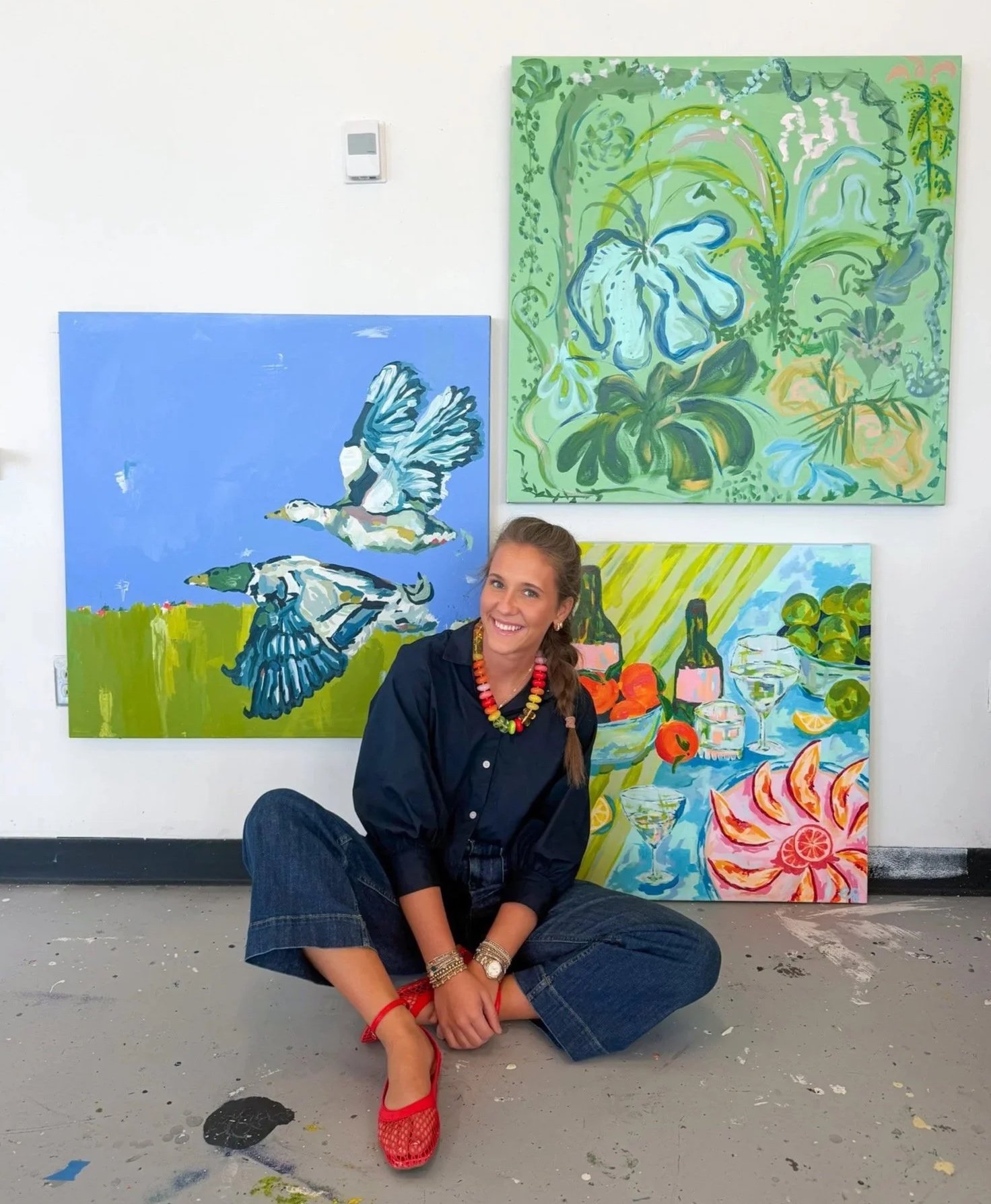 A smiling woman with a long braid, wearing a black shirt, blue jeans, red shoes, and colorful jewelry, seated on the floor in front of a wall with four colorful paintings, including abstract nature scenes and birds.