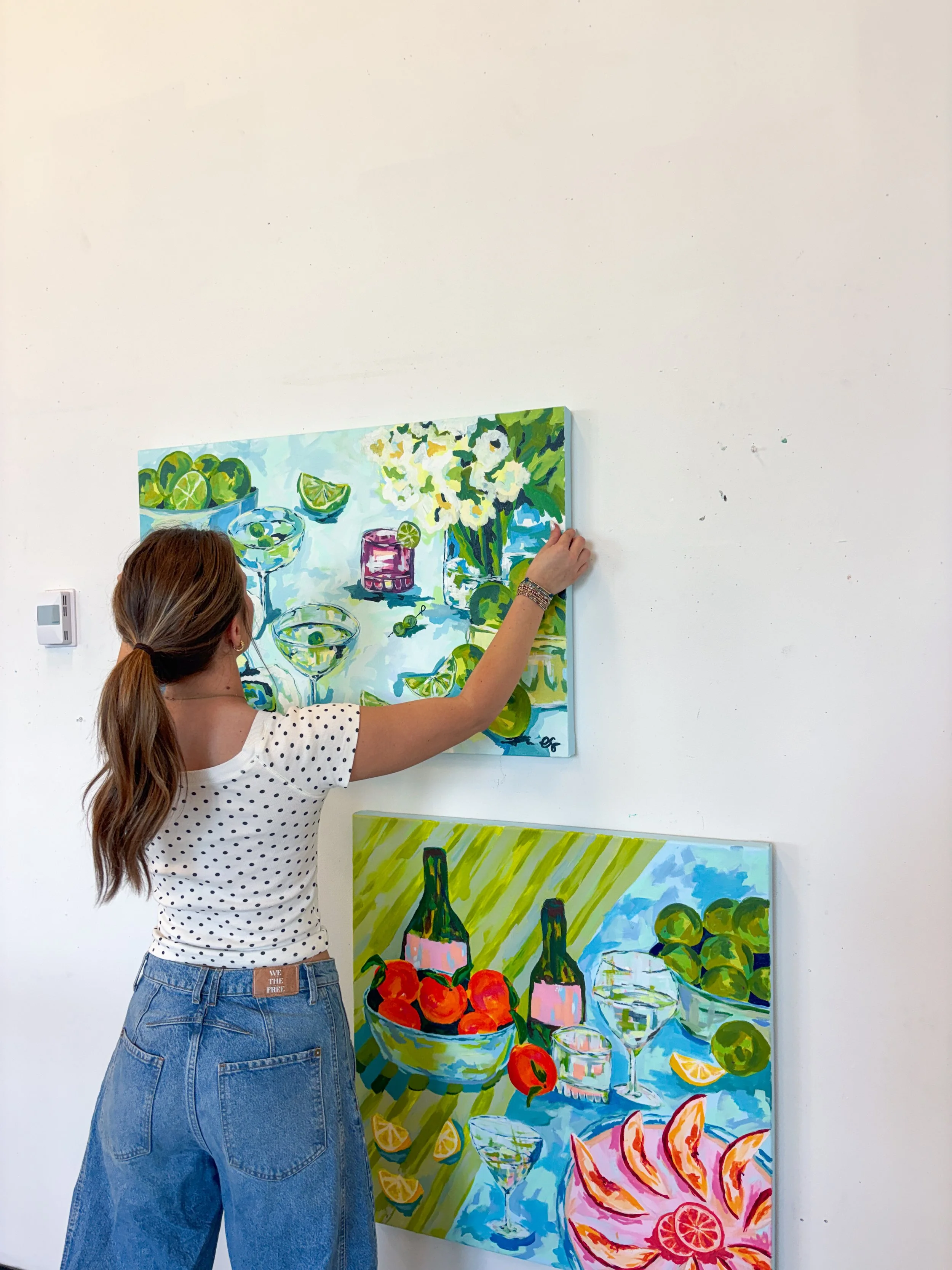 A woman with brown hair in a ponytail, wearing a white shirt with black polka dots and blue jeans, arranging colorful still life paintings on a white wall. The paintings depict drinks, fruits, and eatables with vibrant colors.