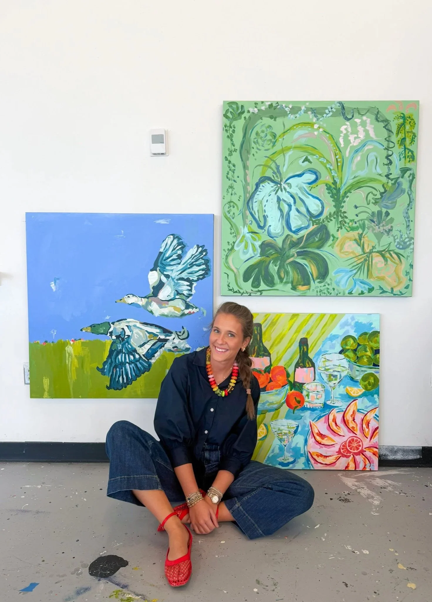 A young woman sitting on the floor in front of colorful paintings featuring birds and flowers. She is smiling, wearing a black shirt, blue jeans, and red shoes, with a beaded necklace and bracelets.