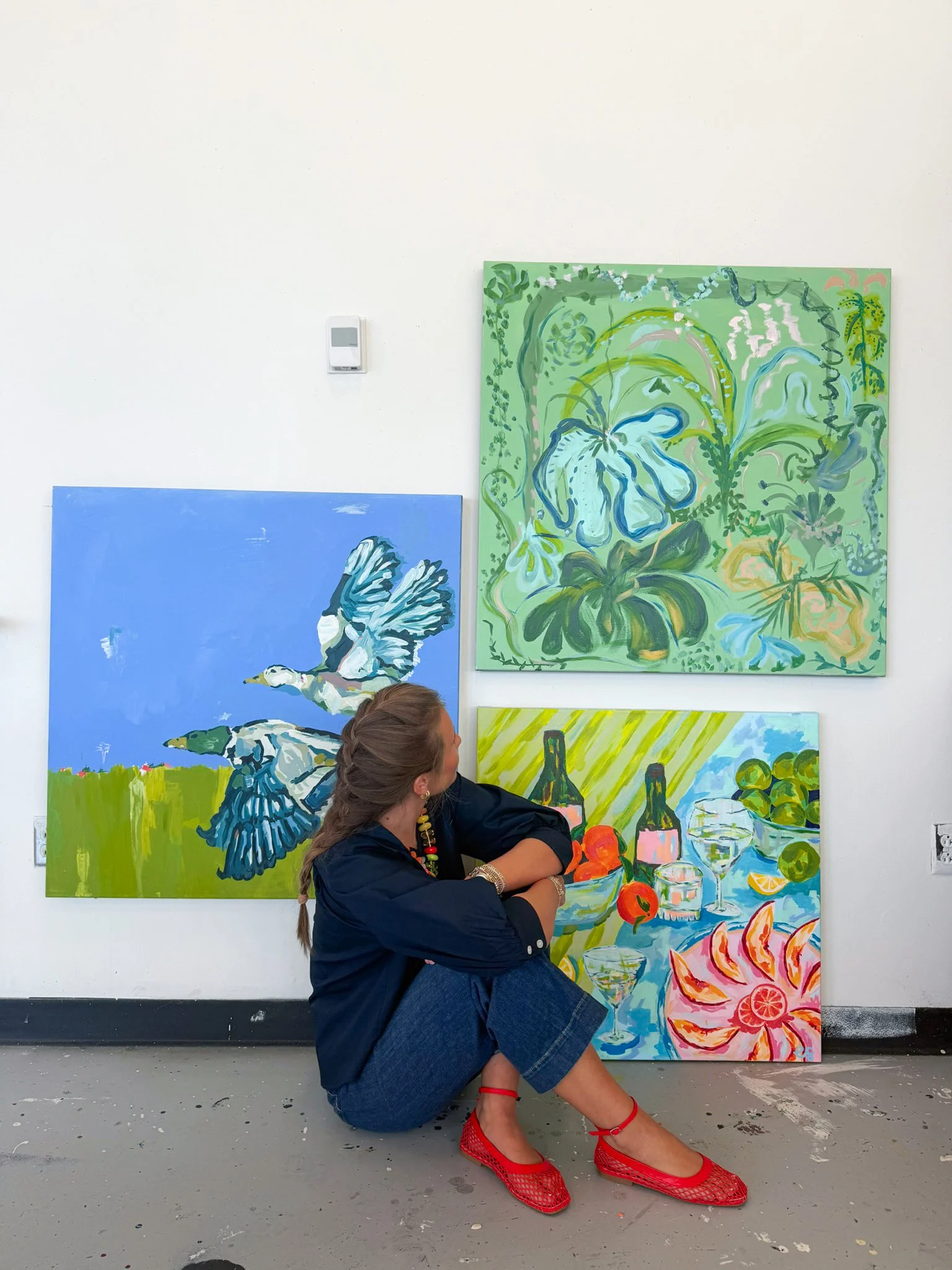 A woman with braided hair wearing a black top and denim shorts sits on the floor in front of colorful paintings, including a blue one with a bird and a green one with a floral design. She is wearing red mesh shoes and has her arms crossed, looking to her right.
