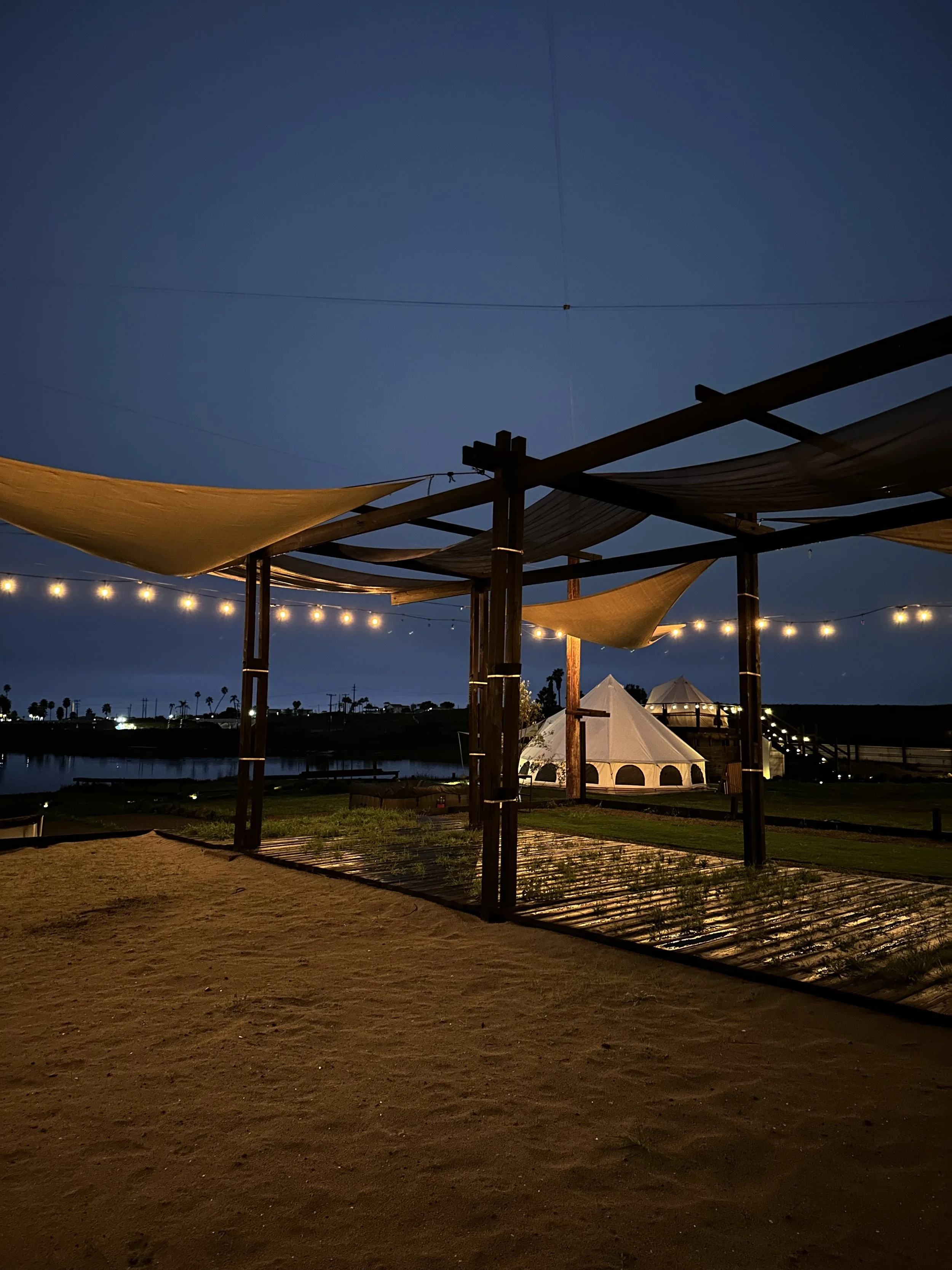 Night view of an outdoor area with string lights hanging overhead, a wooden structure with fabric canopies, and a white tent in the distance near a body of water.