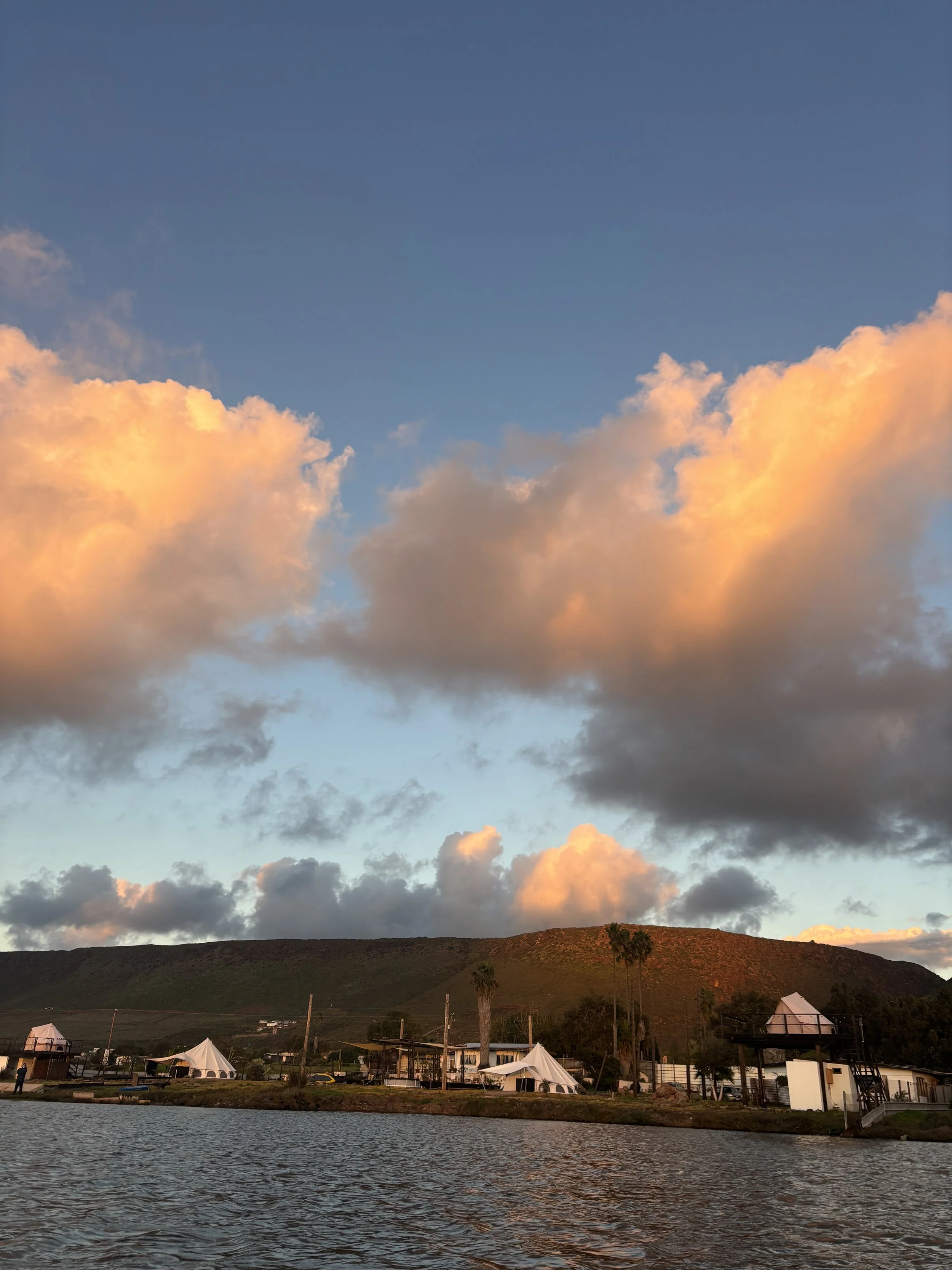 Sunset over a body of water with a mountain in the background, scattered clouds, and structures with tents and palm trees in the foreground.