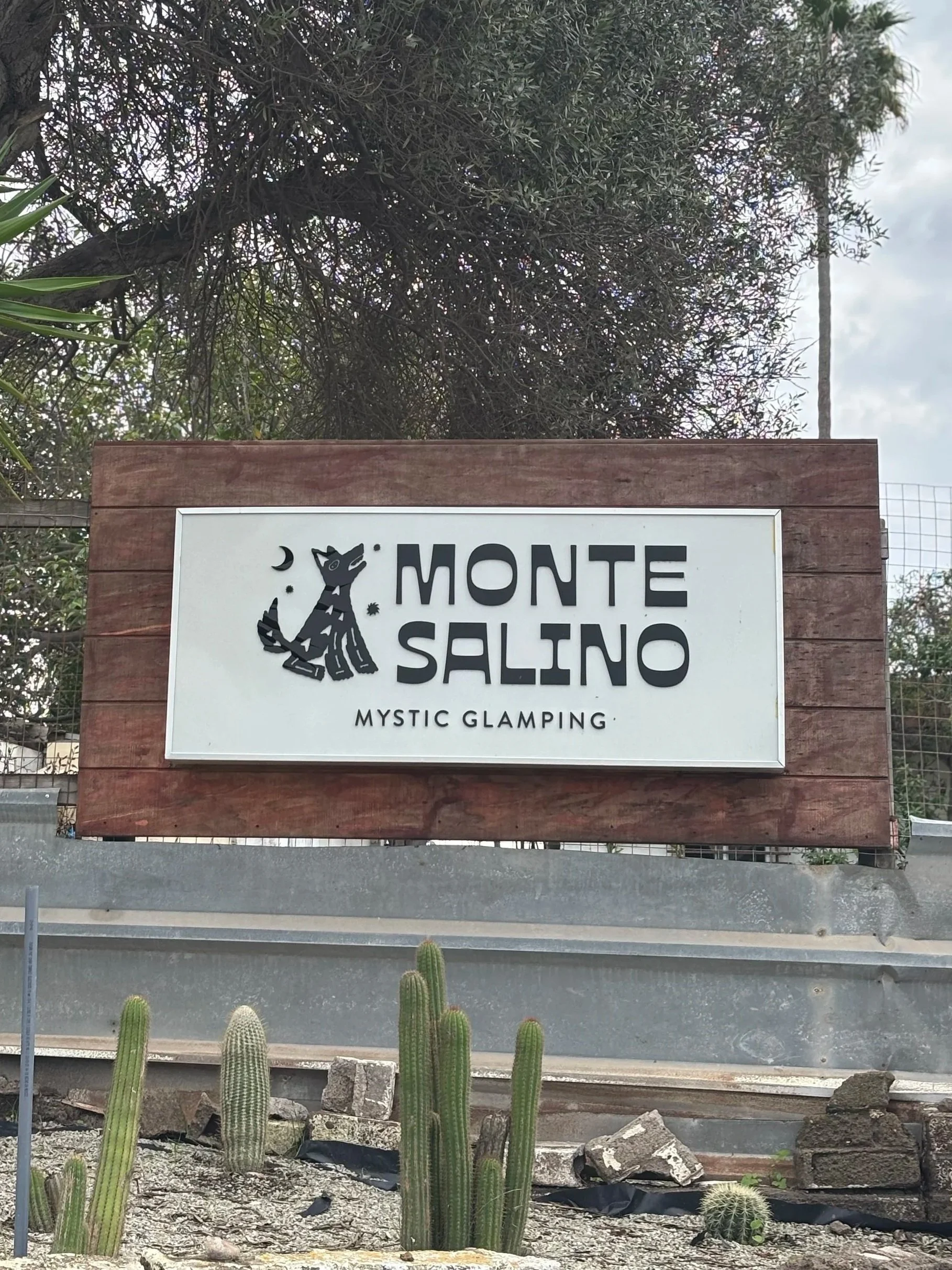 Signboard for Monte Salino Mystic Glamping with a black stylized cat and moon, set against a backdrop of trees and desert plants.