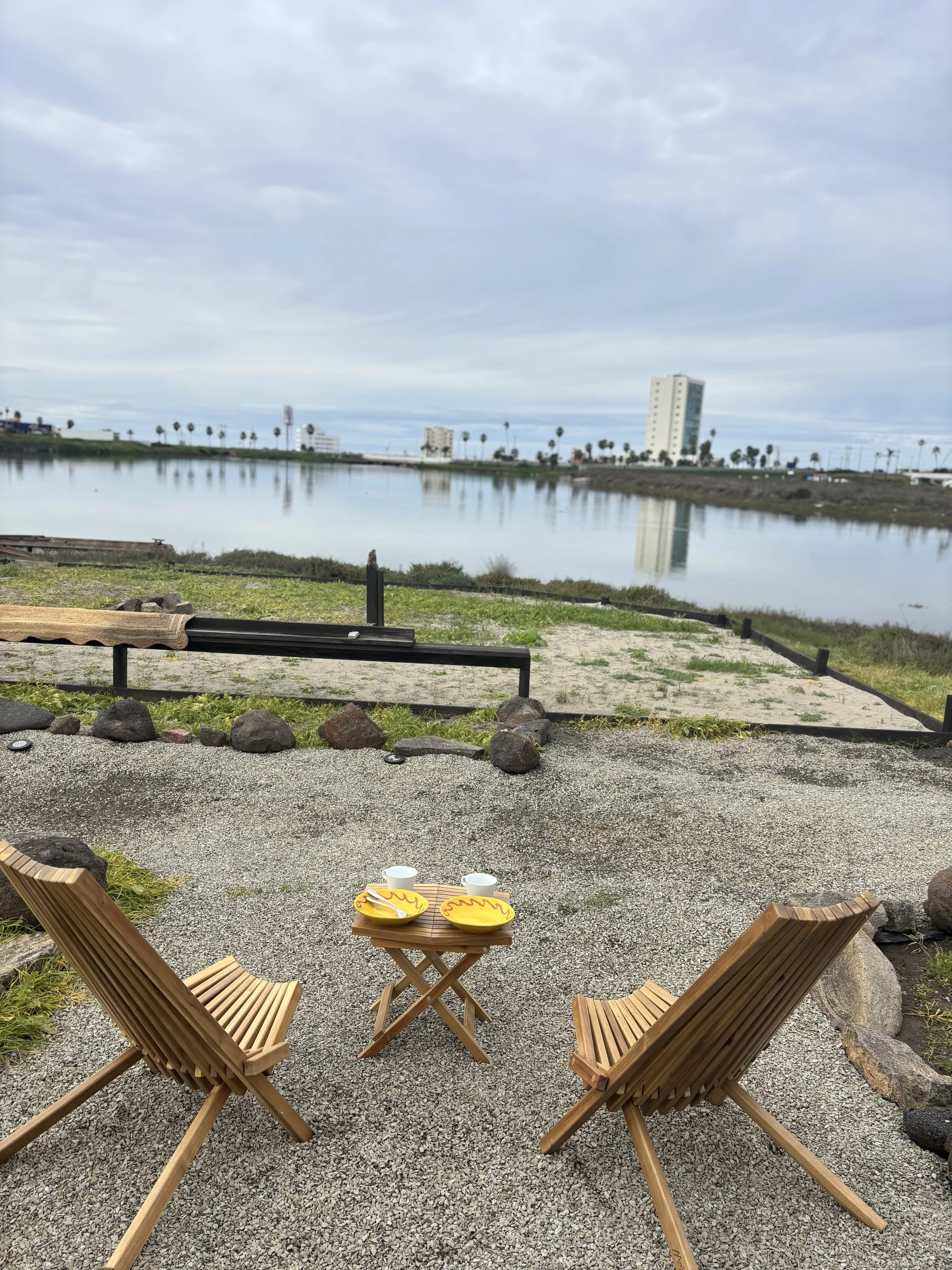 Two wooden chairs and a small wooden table with yellow and white dishes and cups outside near a body of water under a cloudy sky.