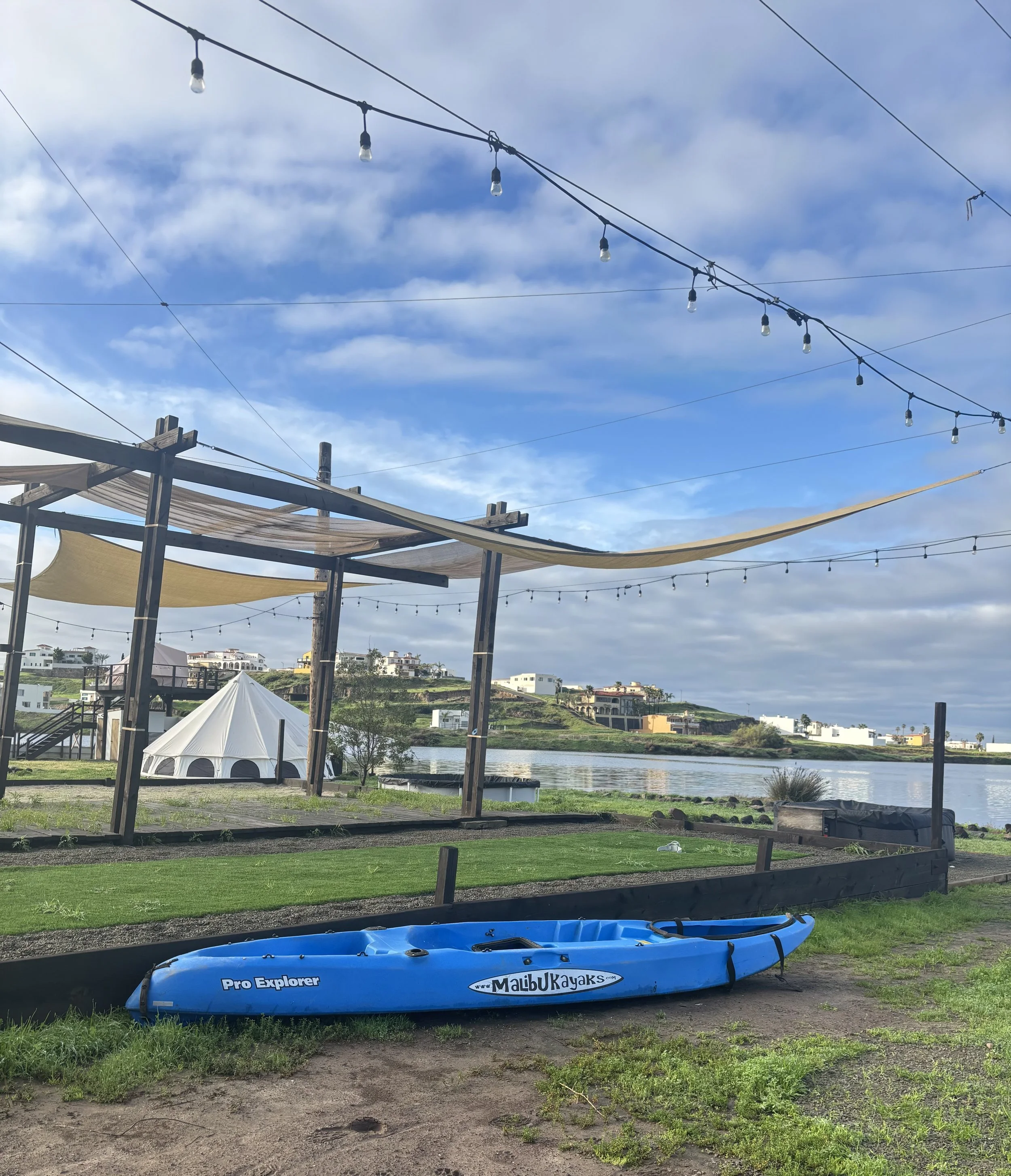 View of a lakeside outdoor area with a blue kayak labeled 'Malibu Kayaks' on the ground, a small white tent, string lights overhead, wooden structures, and houses on a hillside across the water under partly cloudy skies.
