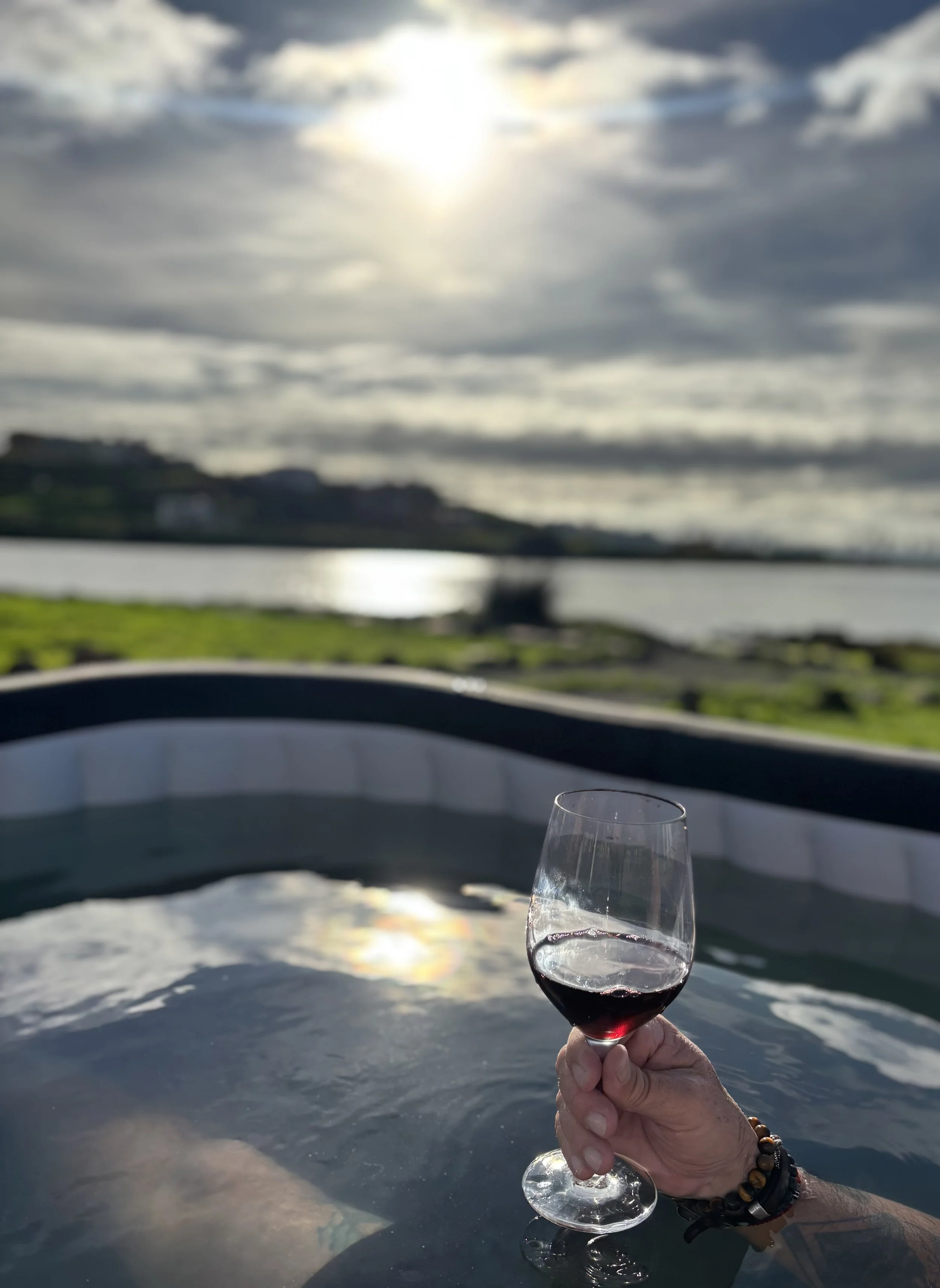 Person holding a glass of red wine in a hot tub overlooking a lake at sunset with cloudy sky.